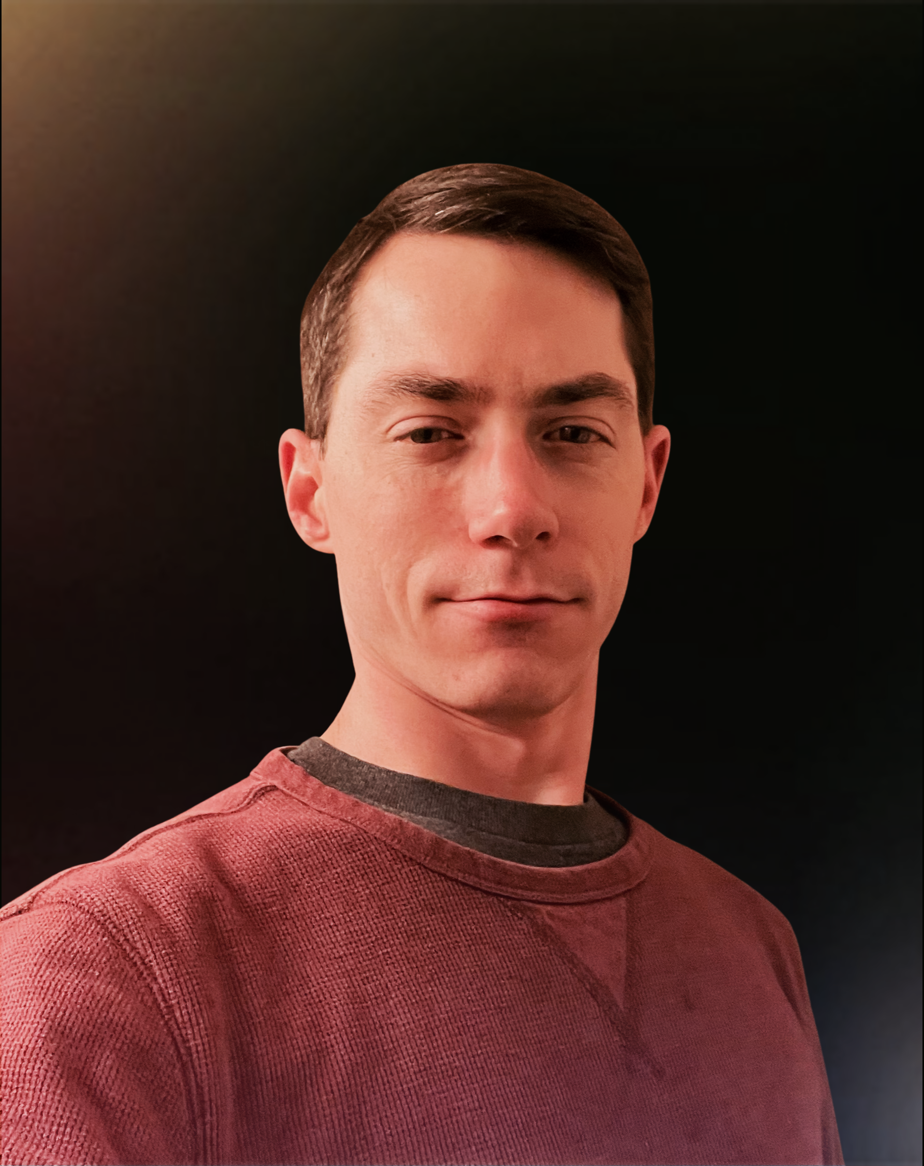 A young man with short brown hair and light skin is wearing a red textured sweater with a dark collar. He is smiling slightly and is looking toward the camera. The background is black.
