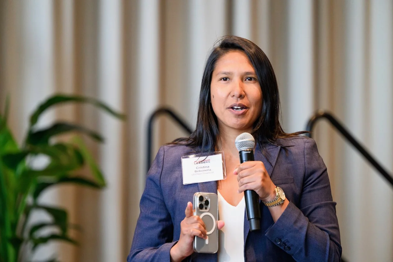 Woman speaking at a conference, holding a microphone and a smartphone, wearing a name badge and business attire, standing in front of a curtain with a plant nearby.