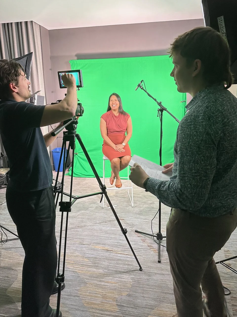 A woman in a red dress is sitting on a stool in front of a green screen, smiling during a video shoot. Two men are operating camera equipment; one is adjusting the camera, and the other is holding a script or notes, with a microphone set up nearby.
