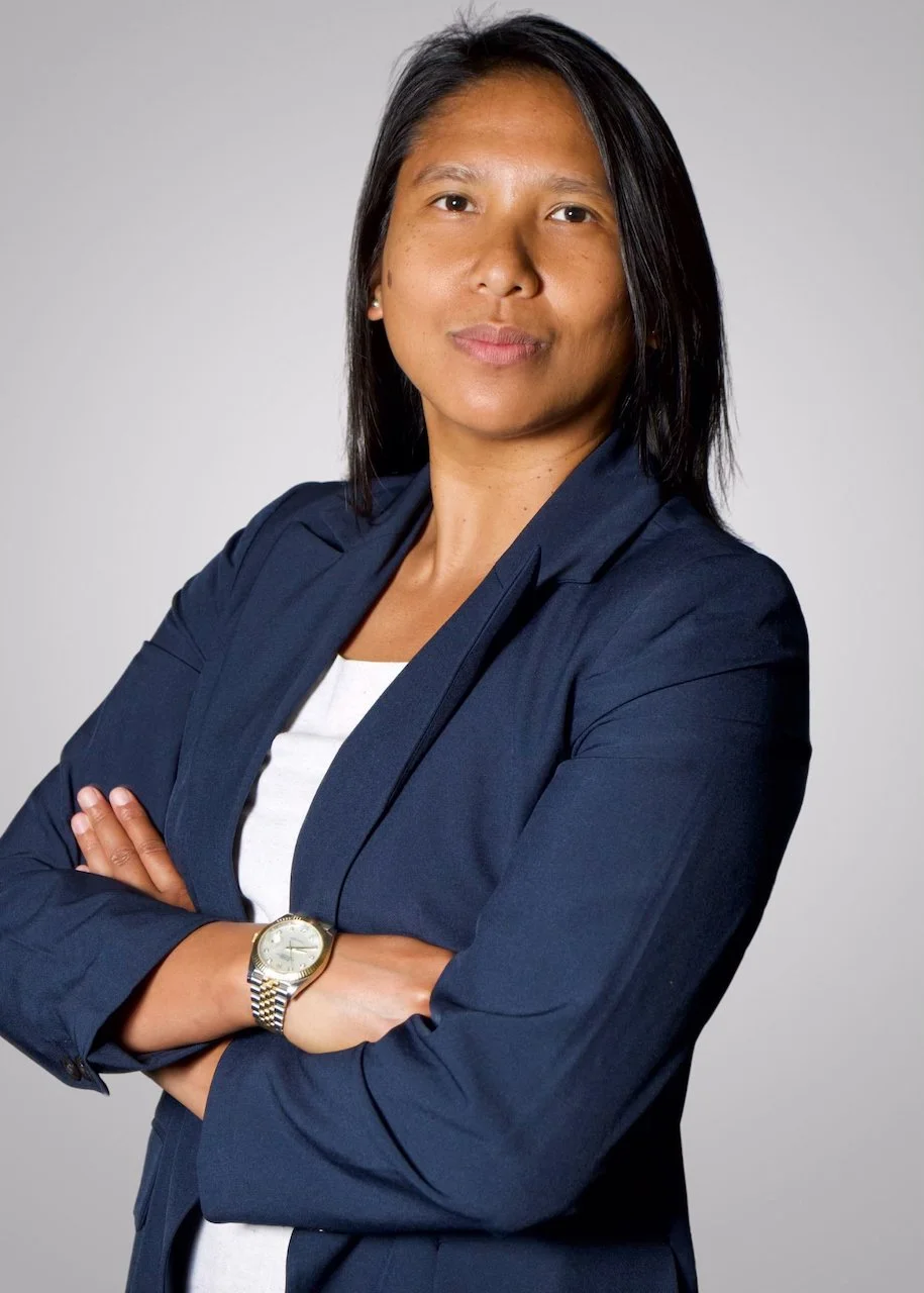 Professional woman with black hair, wearing a navy blazer and white shirt, crossing arms, silver watch, neutral background.