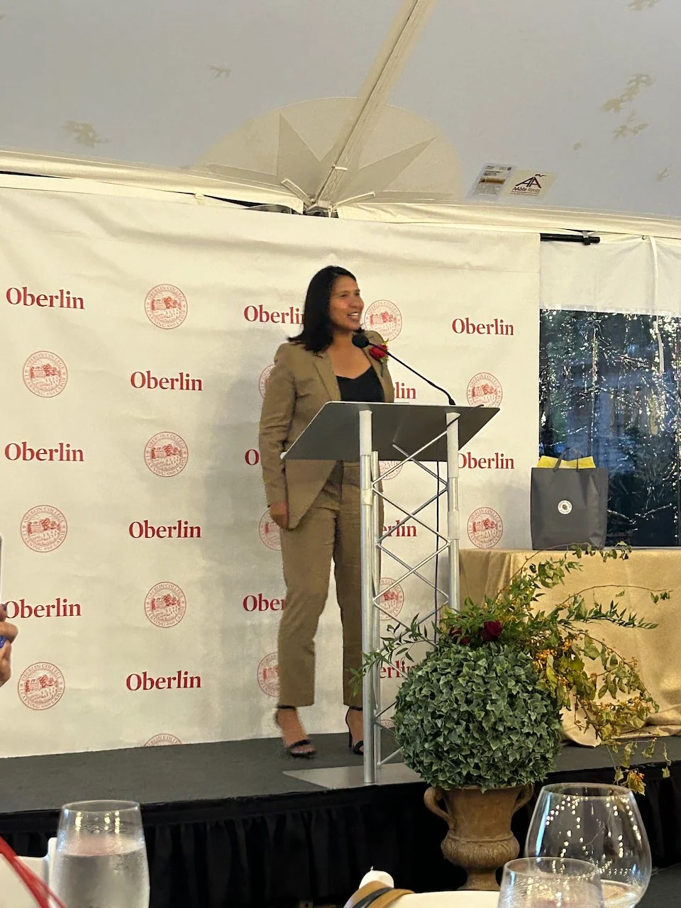 A woman in a tan suit speaking at a podium during an event with Oberlin repeated on the backdrop.