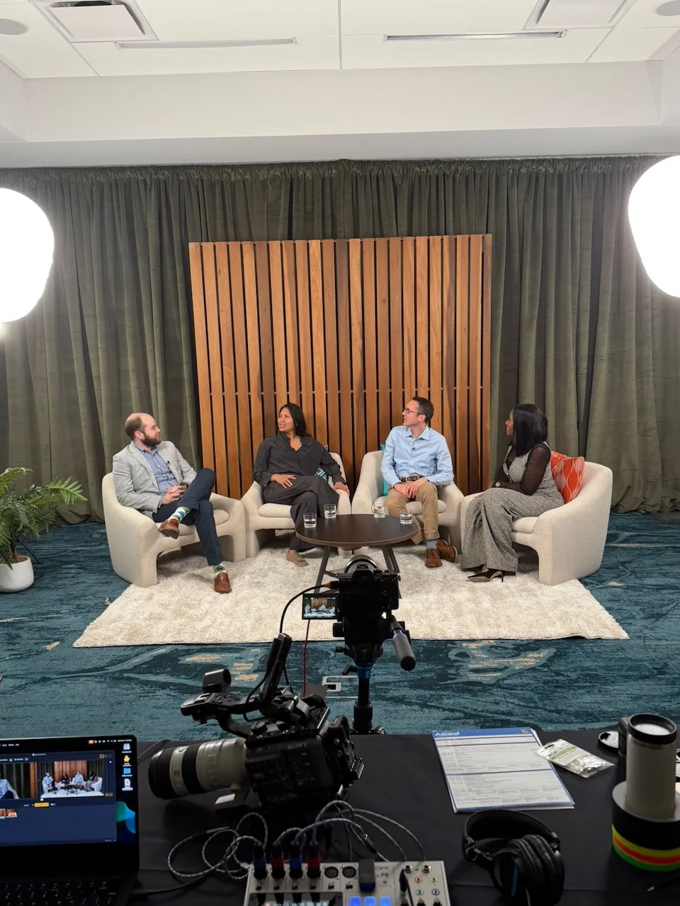 Four people sitting on chairs in a studio setting, engaged in conversation. Two large lights illuminate the scene, with a camera and equipment facing them. The background features a dark curtain and a wooden panel, with a white rug under the chairs. There is also a small table with glasses and equipment on it.