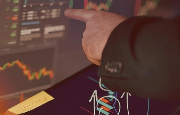 A person in a business suit points at a computer screen displaying stock market charts and graphs, with a sticky note in the foreground.