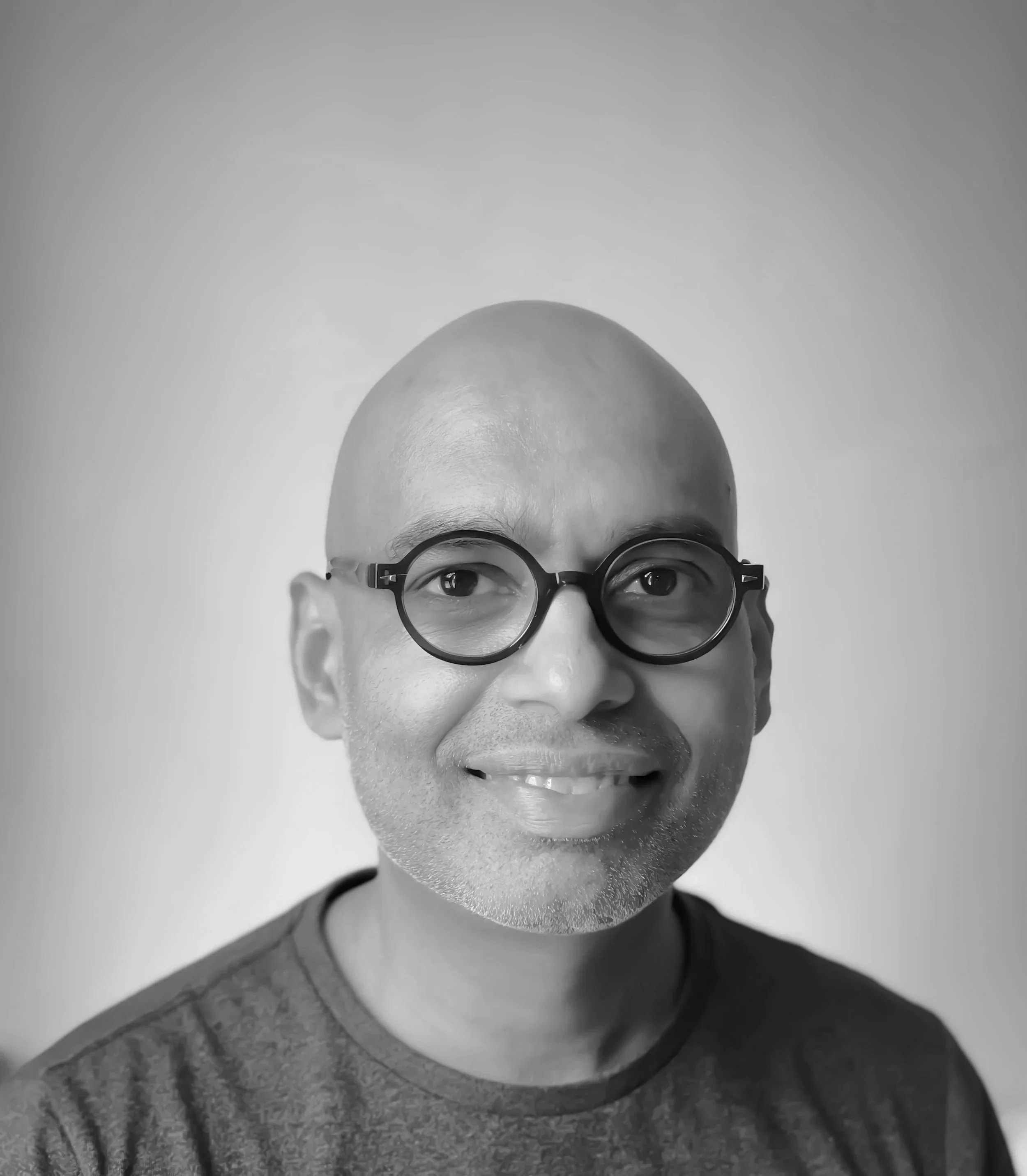 Black and white photograph of Neil Salisbury, with glasses, smiling, wearing a jacket with a textured fabric, with a plain wall background.