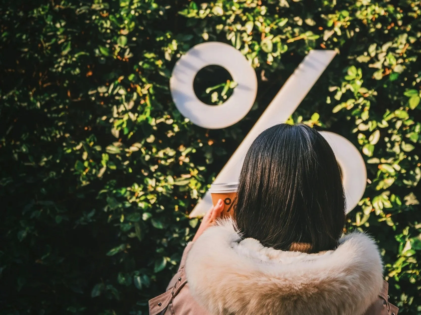 A woman with dark hair, wearing a beige coat with a fur collar, holding a coffee cup, stands in front of a green leafy hedge with large white percentage sign on it.