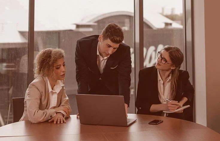 Three business professionals in a meeting room, two women and one man, looking at a laptop screen on the table, with notes and a smartphone in view.