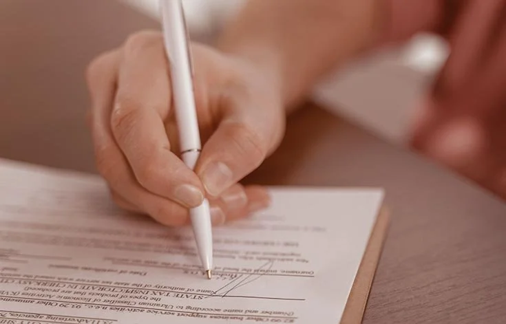 Close-up of a person's hand signing a document with a white pen on a clipboard.