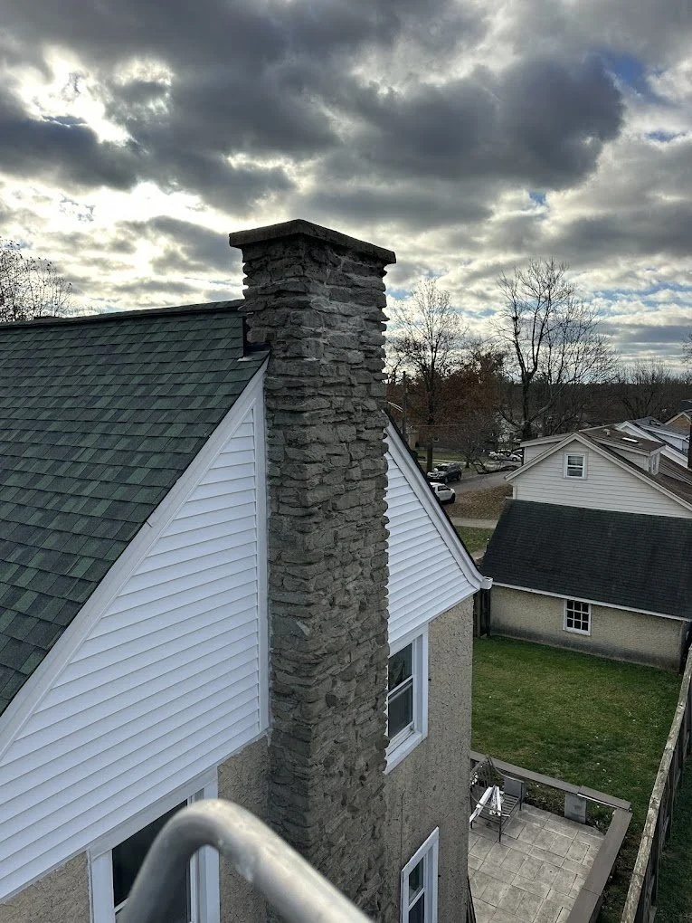 A house with a stone chimney, white siding, green shingles on the roof, and a small backyard patio with chairs, under a cloudy sky.