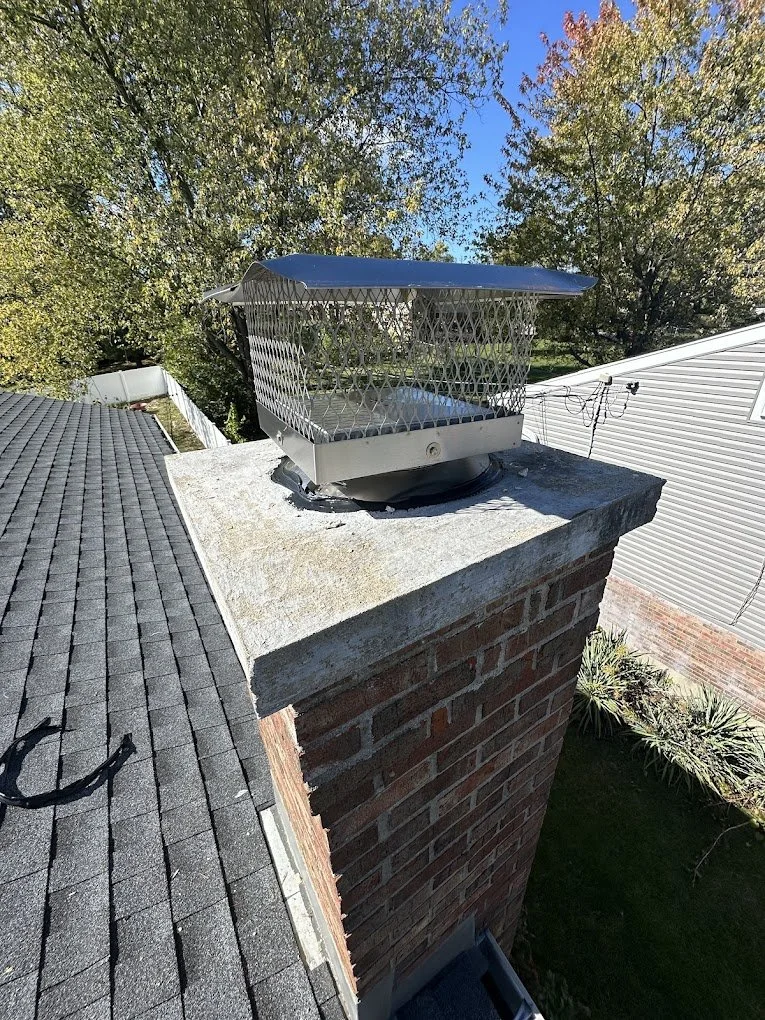 View of a brick chimney with a metal chimney cap on top. The cap has a protective cage around it, with a sloped roof. The chimney is on a rooftop with asphalt shingles, and a clear blue sky with some trees is visible in the background.
