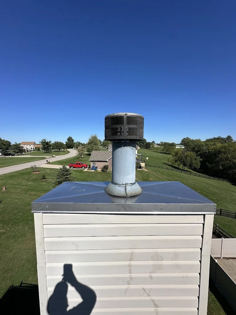 Close-up of a house chimney with a black vent cap on a metal roof, in a suburban neighborhood with houses, trees, and a red truck visible in the background on a clear, sunny day.
