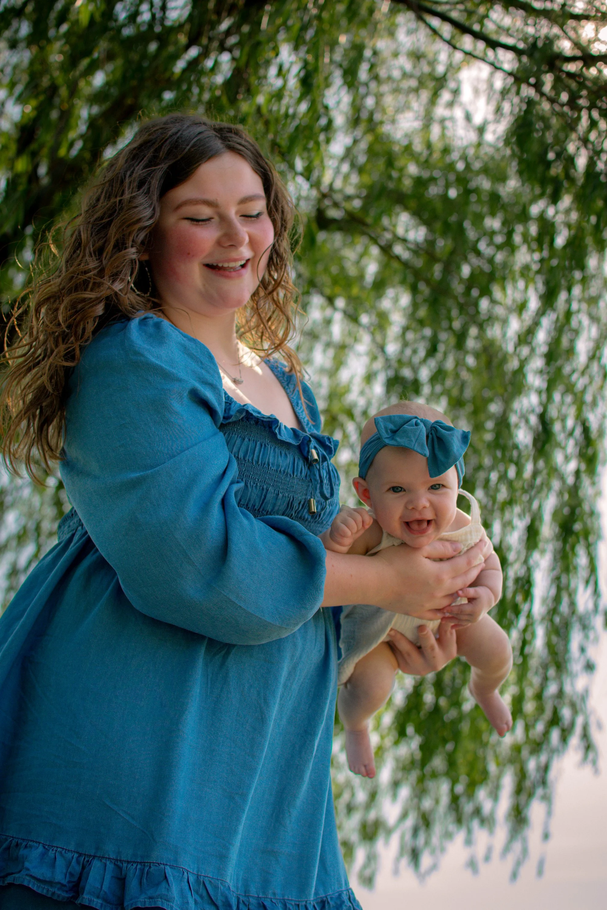 Mother holding her smiling baby beneath a willow tree during an outdoor portrait session.
