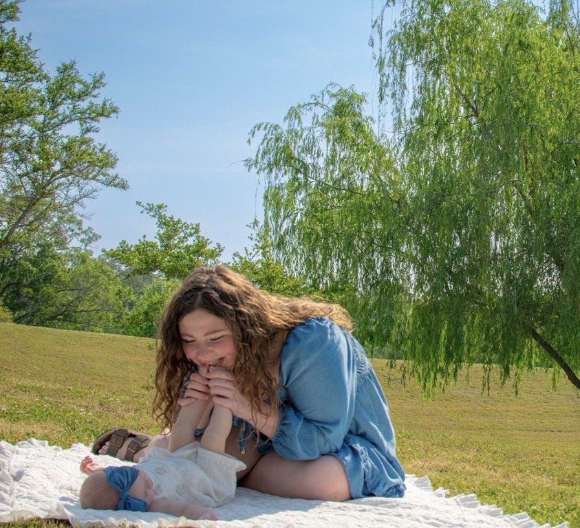 Mother smiling while playing with her baby on a blanket during an outdoor lifestyle session.