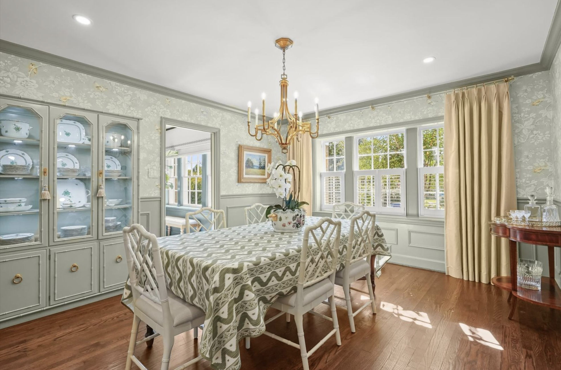A dining room with a wooden table covered by a patterned tablecloth and surrounded by white chairs. There is a china cabinet filled with dishes and a floral painting on the wall. The room has large windows with blinds and beige curtains, a chandelier hanging from the ceiling, and hardwood floors.