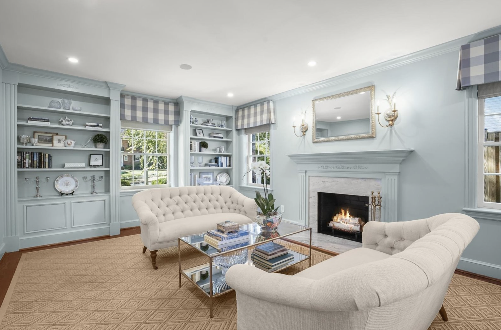 A cozy living room with light blue walls, built-in shelves, a fireplace, cream-colored tufted sofas, a glass coffee table, and windows with blue checkered valances.