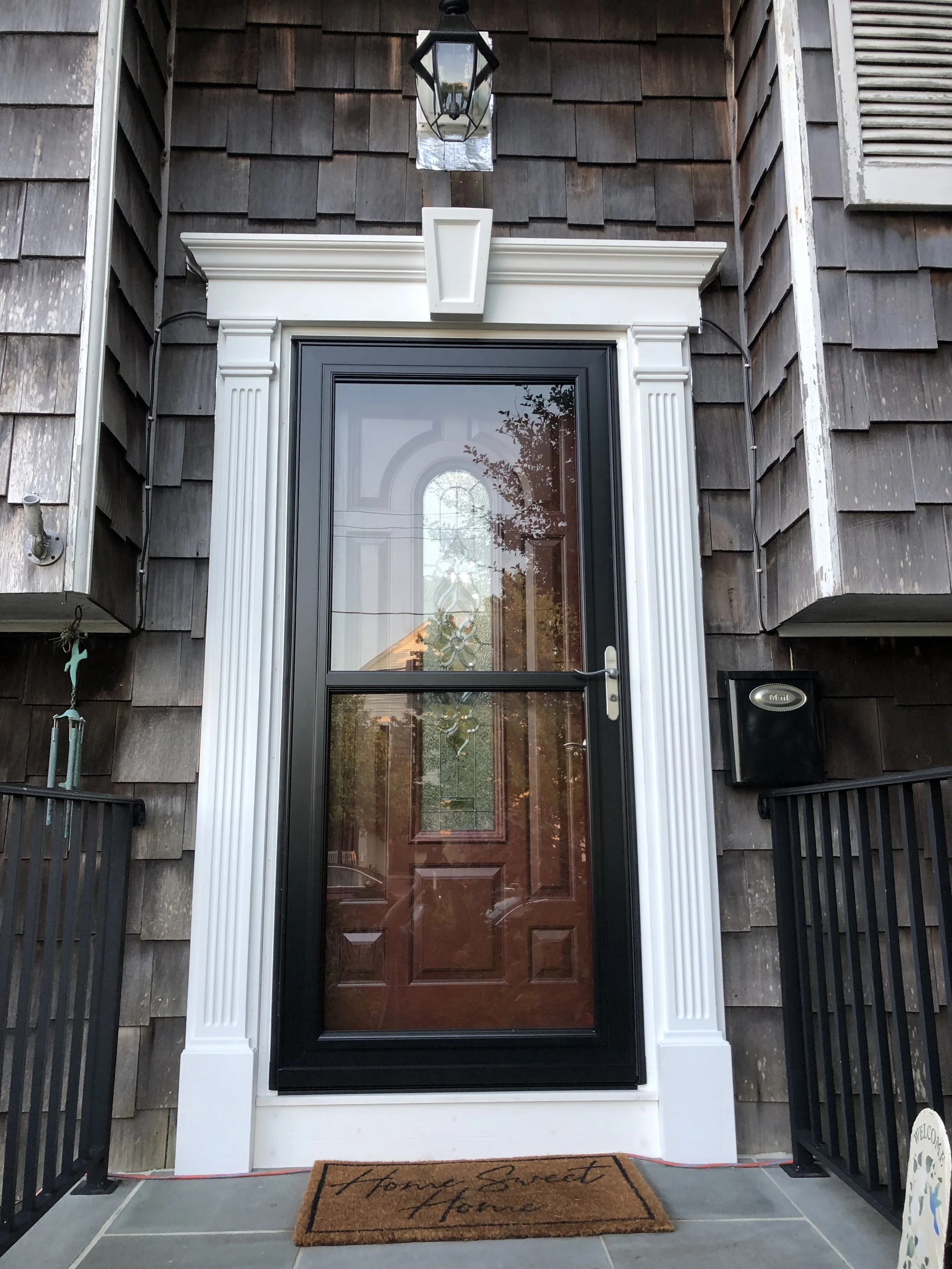 Front entry door with a brown wooden door, white trim, and a black metal frame, surrounded by dark brown wooden shingles on the house exterior. A black lantern-style light fixture hangs above the door, and a black mail slot is mounted on the wall to 