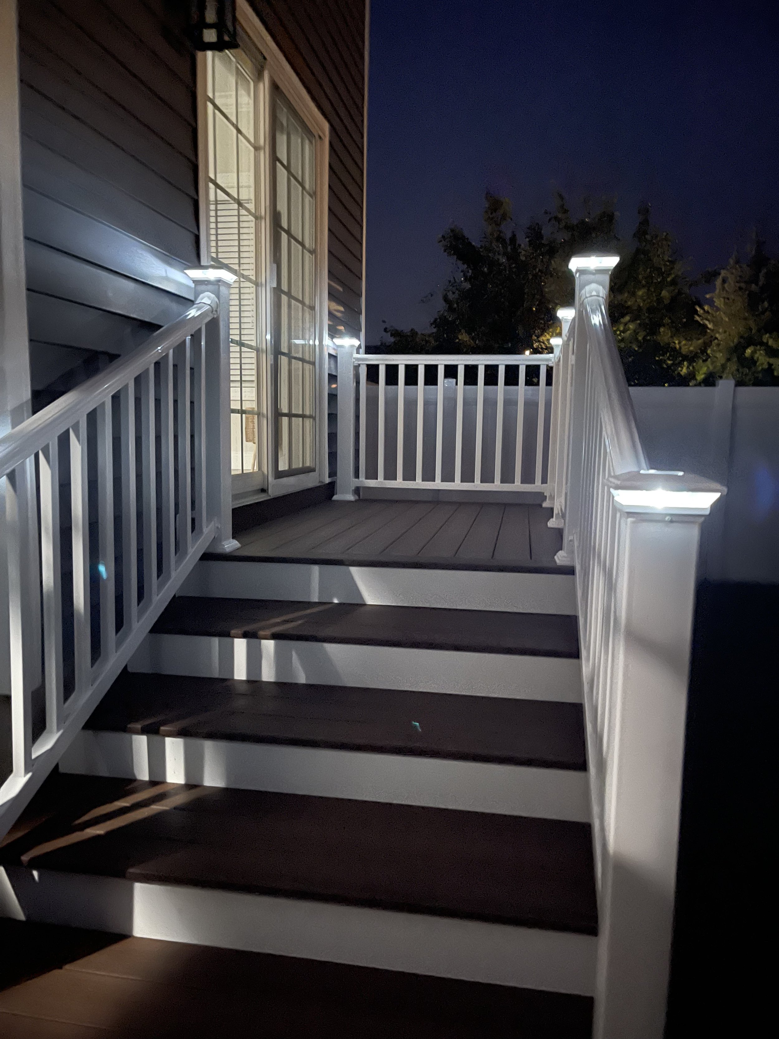A wooden staircase with white railing leading to a sliding glass door on a house exterior. The house siding is dark brown, and there is a black outdoor wall lantern above the door. It is nighttime.