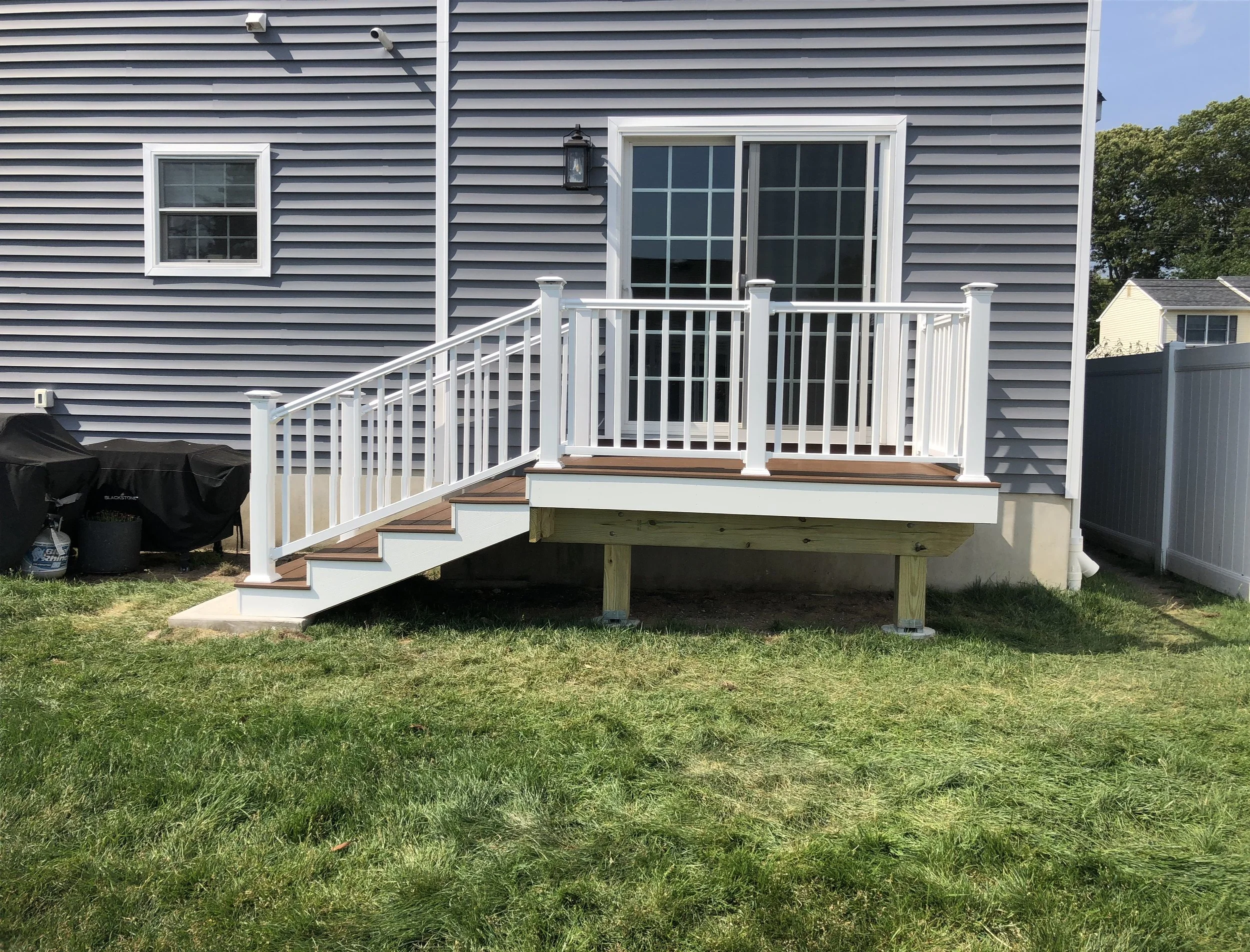 Backyard deck with white railing and sliding glass door, gray house siding, small window, outdoor light fixture, and a grassy yard with trees in the background.