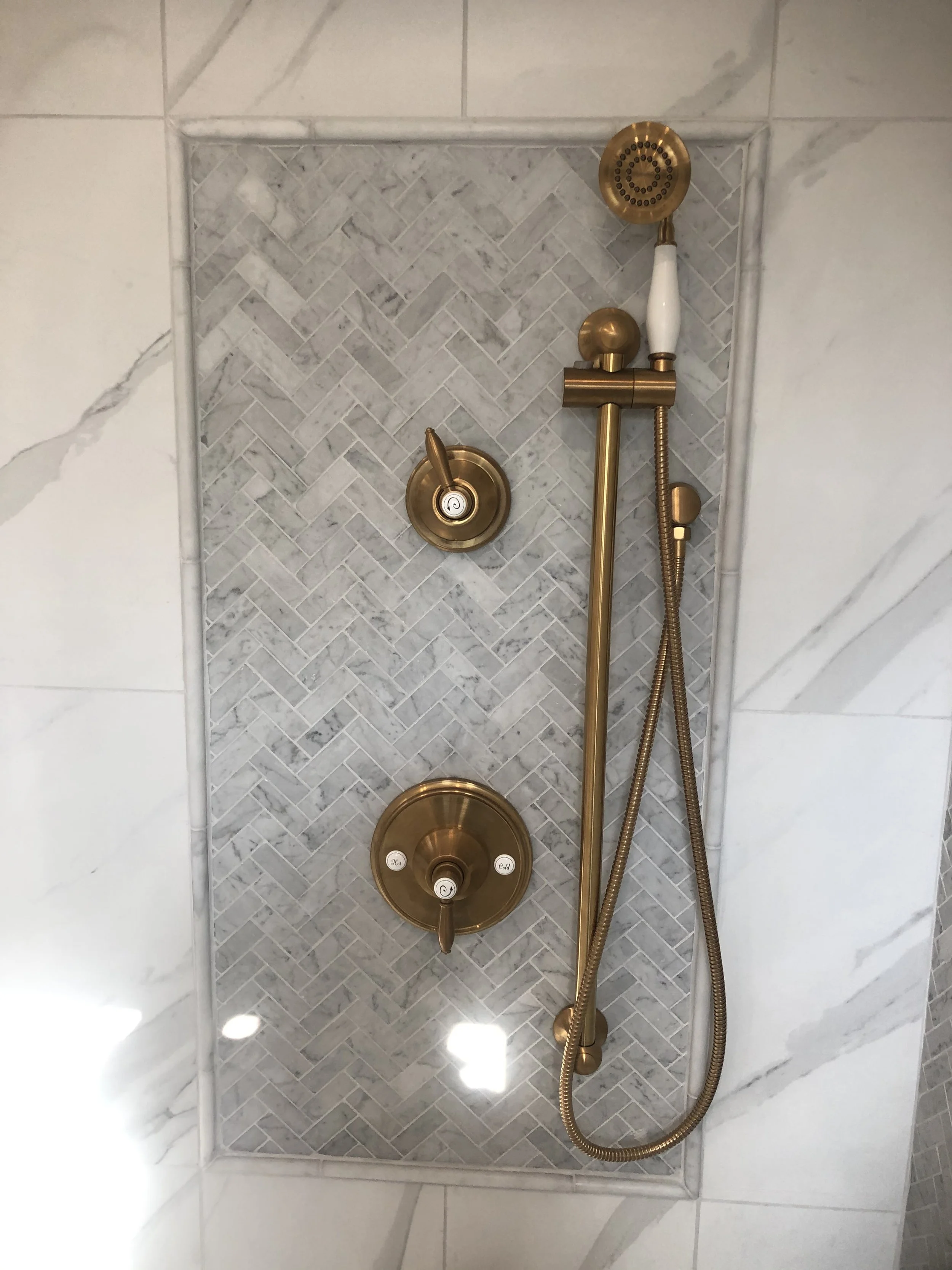 A shower with a gold shower head, controls, and a hose, set against a tiled wall with a herringbone pattern in a bathroom.