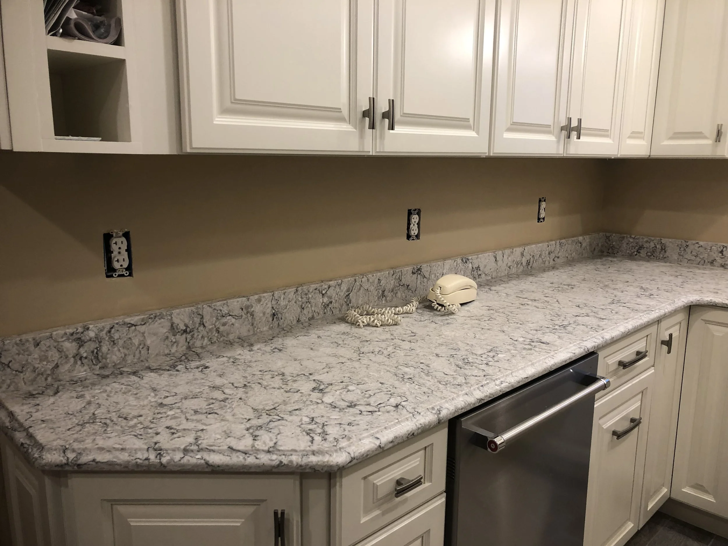 A kitchen countertop with a marbled pattern, an old-fashioned beige telephone, and several empty electrical outlets. Above the countertop are white cabinets with metallic handles.