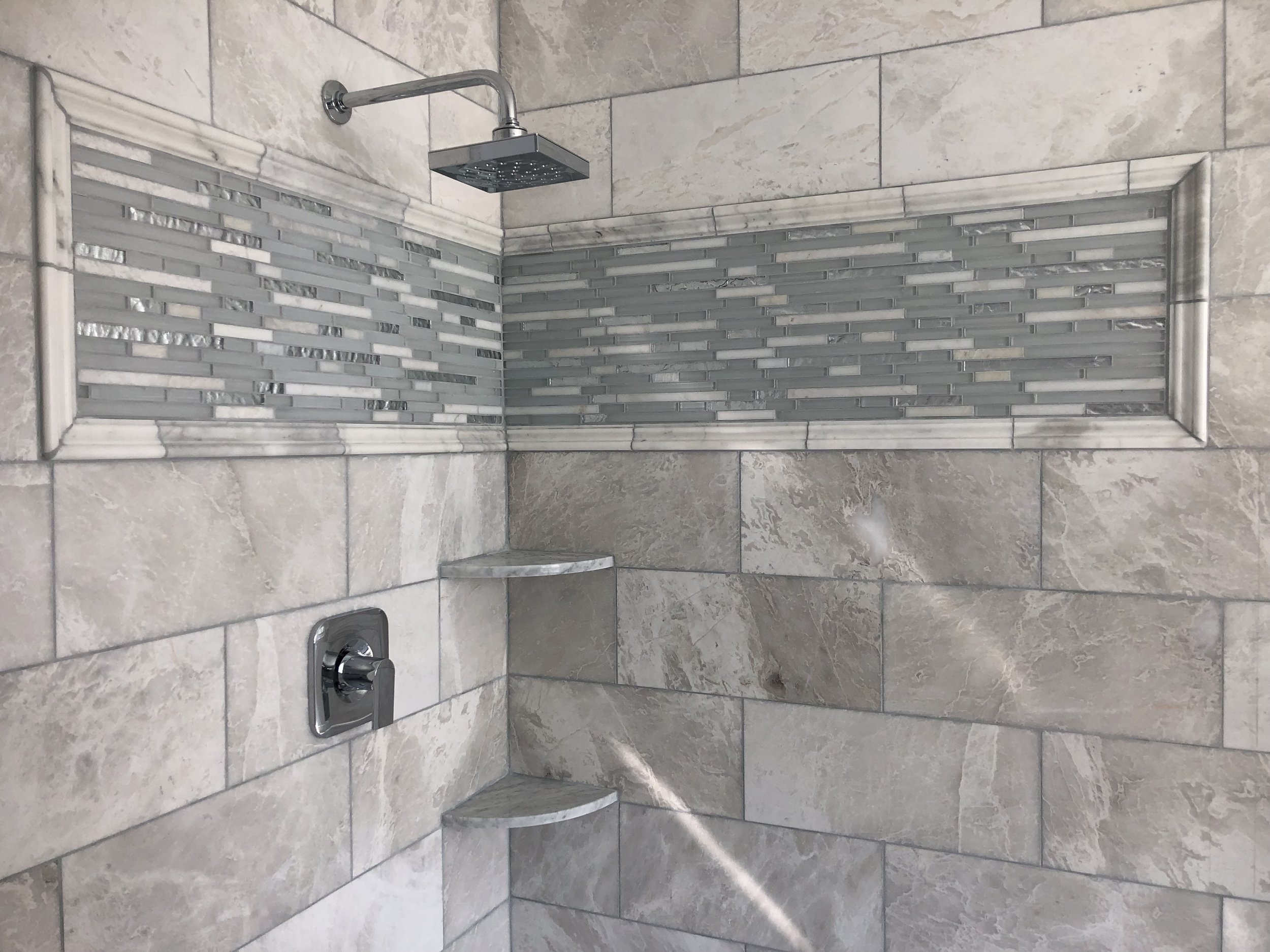 A shower stall with beige tiles on the walls and a decorative strip of smaller, rectangular glass tiles. There is a square showerhead attached to the wall, a control handle, and a built-in corner shelf.