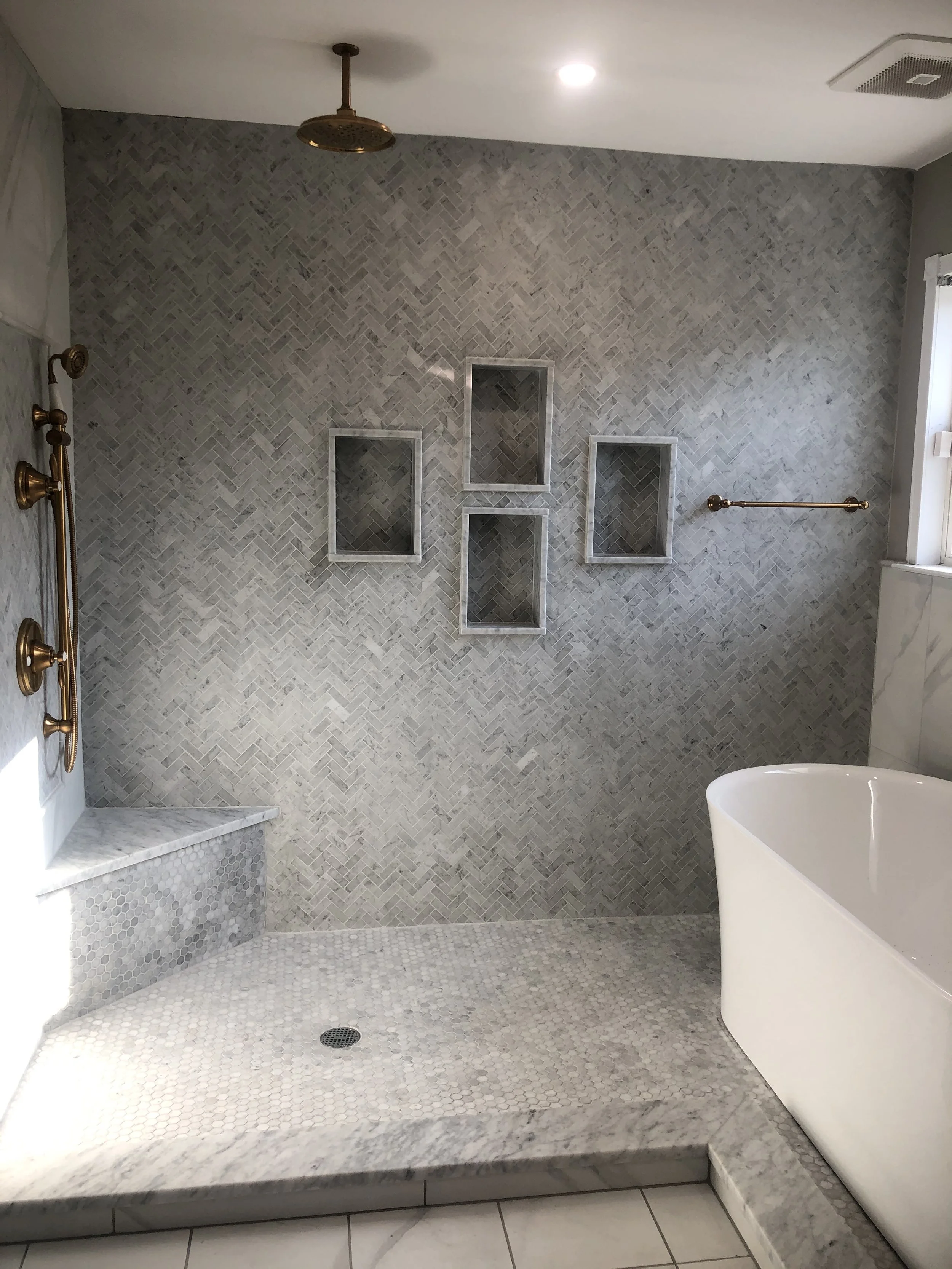 Modern walk-in shower with corner shelves, marble and tile surfaces, a rainfall showerhead, a handheld shower, and a partial view of a bathtub to the right.