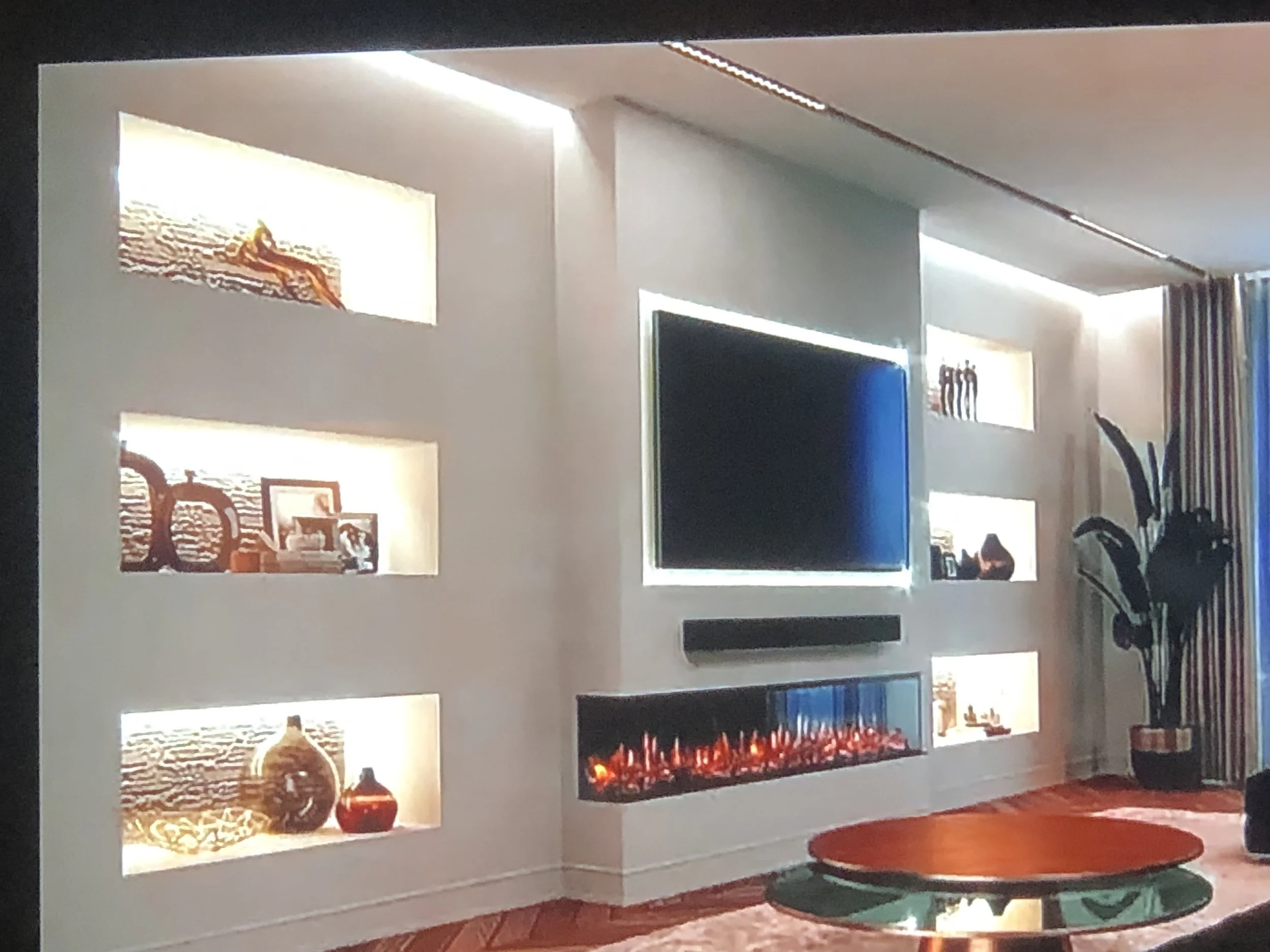Living room with built-in wall unit featuring open shelves displaying decorations and photos, a wall-mounted flat-screen TV, a fireplace with an orange glow, a striped curtain, and a partial view of a person sitting near the TV.