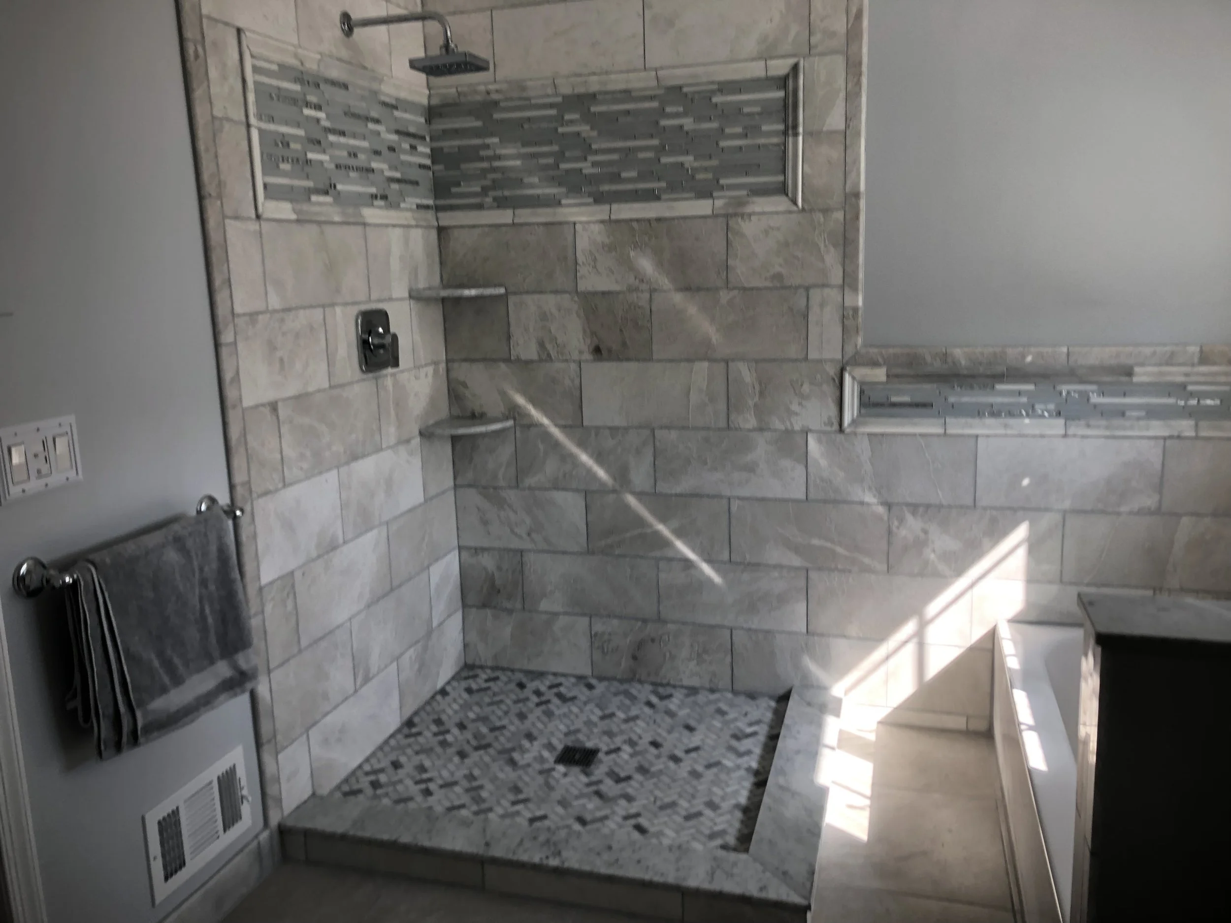 A bathroom shower area with a built-in shelf, a rainfall showerhead, and tiled walls. There is a towel rack with a gray towel on the left side and a patterned floor mat on the floor.