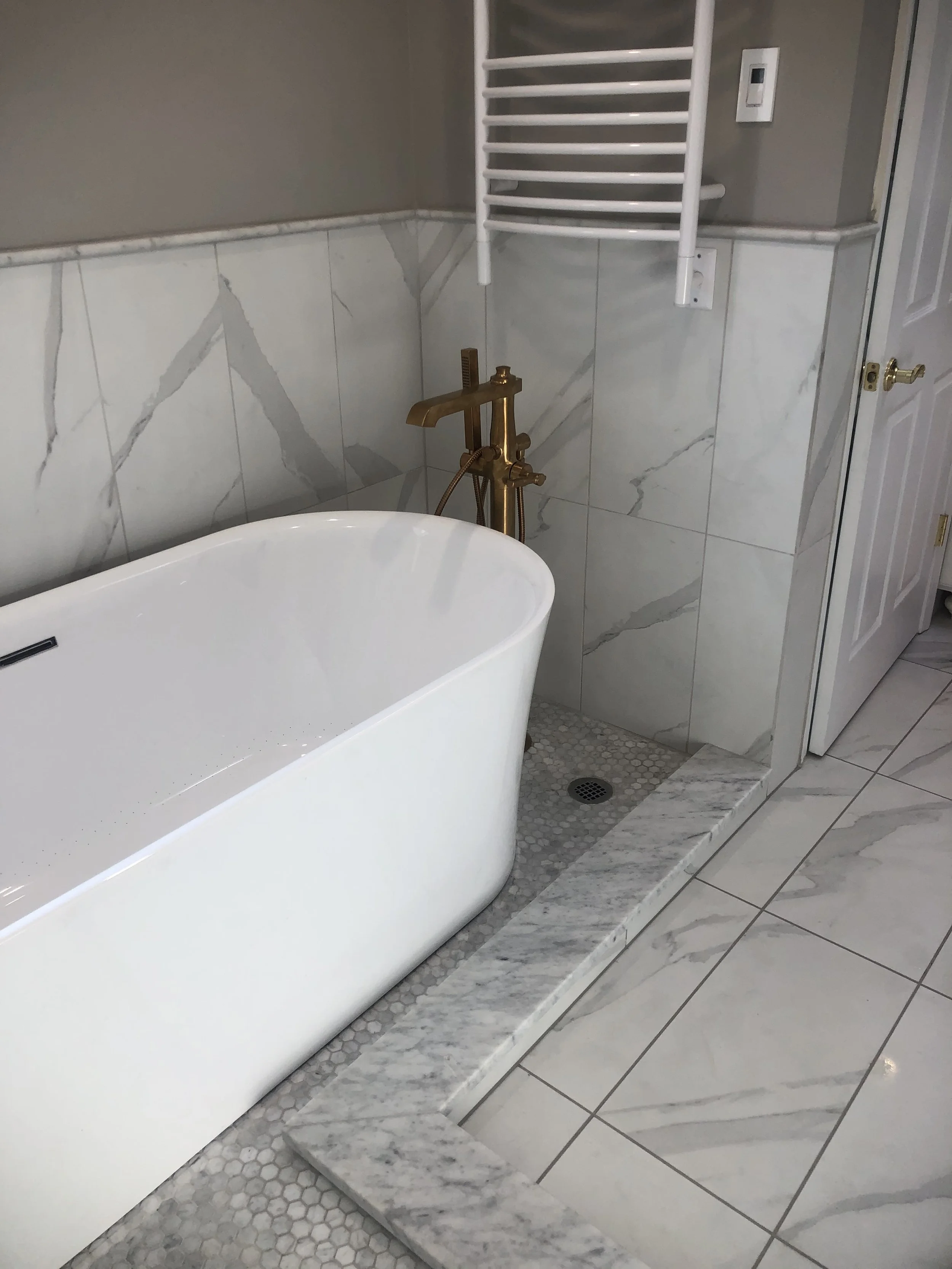 Close-up of a bathroom corner showing a white bathtub, a bronze faucet, marble tiles, a heated towel rack, and a control switch on the wall.