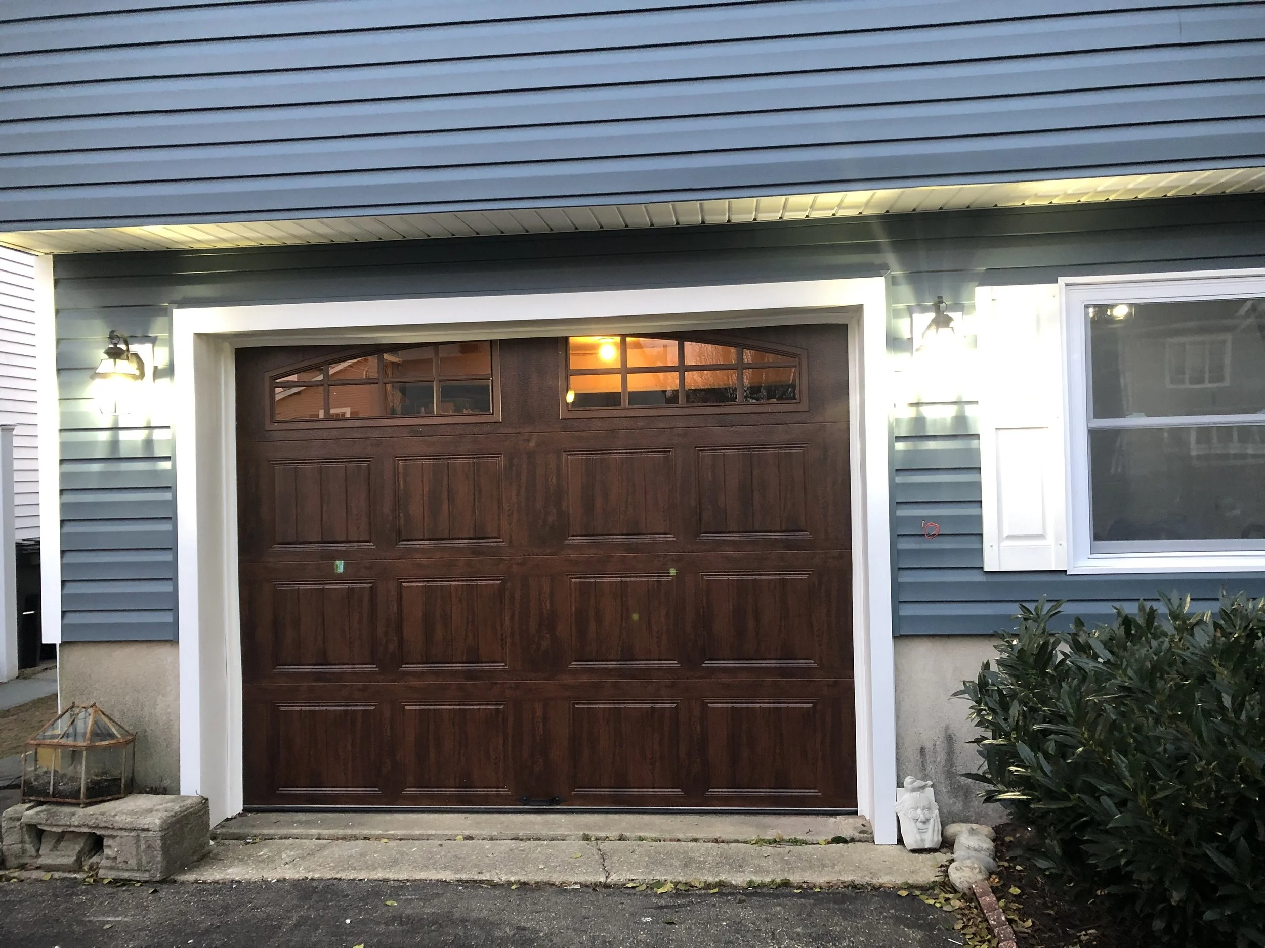 Garage door with two small windows at the top, surrounded by light green siding, illuminated by exterior lights on each side, plant and garden statue on the right.