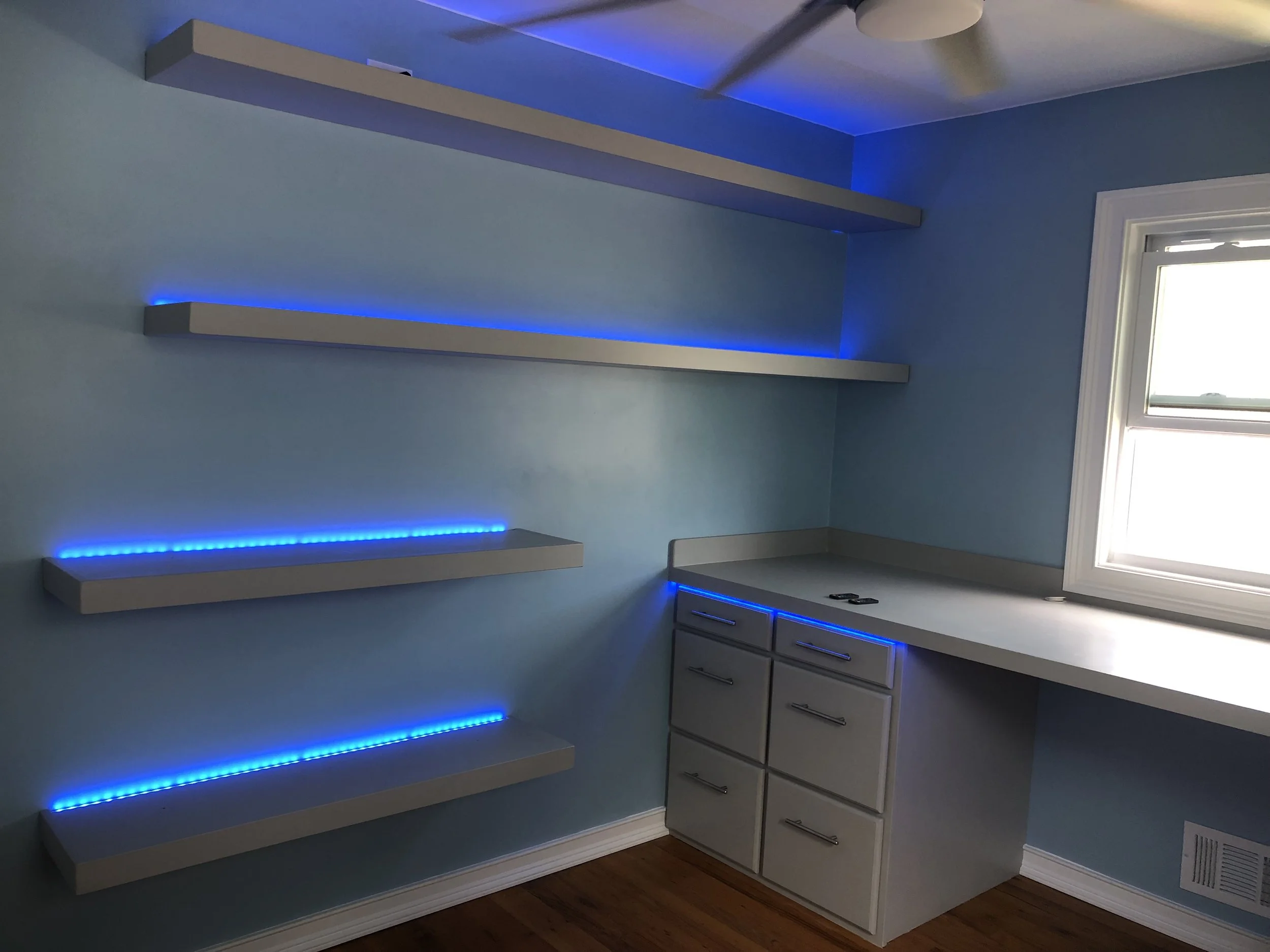 Room with light blue walls, three floating shelves with blue LED lighting, a window, and gray cabinets with black handles.