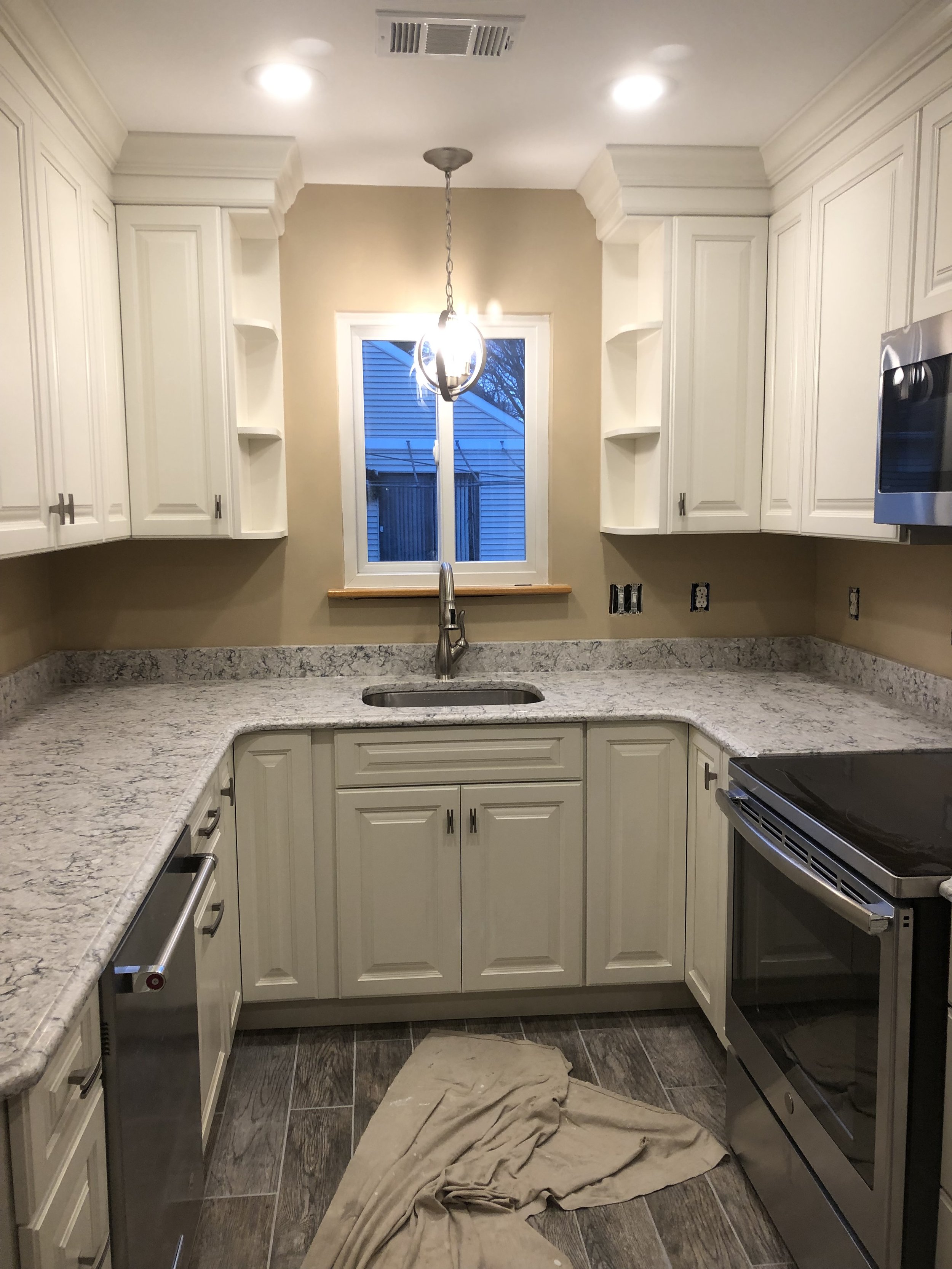 Unfinished kitchen with white cabinets, granite countertops, a window above the sink, a stove, and a chandelier. There is a beige cloth on the floor.