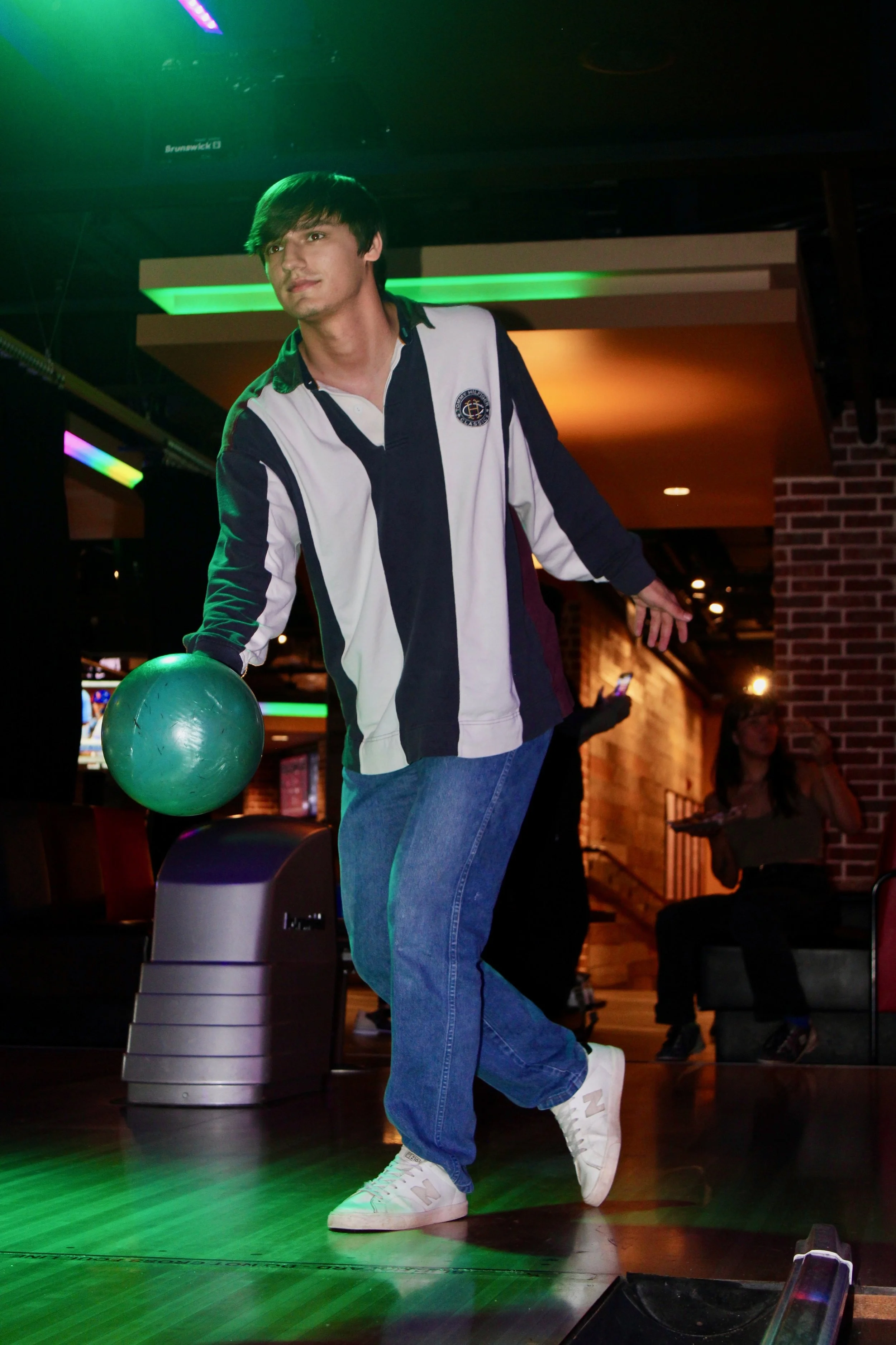 A young man in a black, white, and dark blue long-sleeve shirt, blue jeans, and white sneakers is bowling. He is holding a green bowling ball and appears to be preparing to roll it down the lane. The setting is a bowling alley with dim lighting, colored lights, and other people in the background.