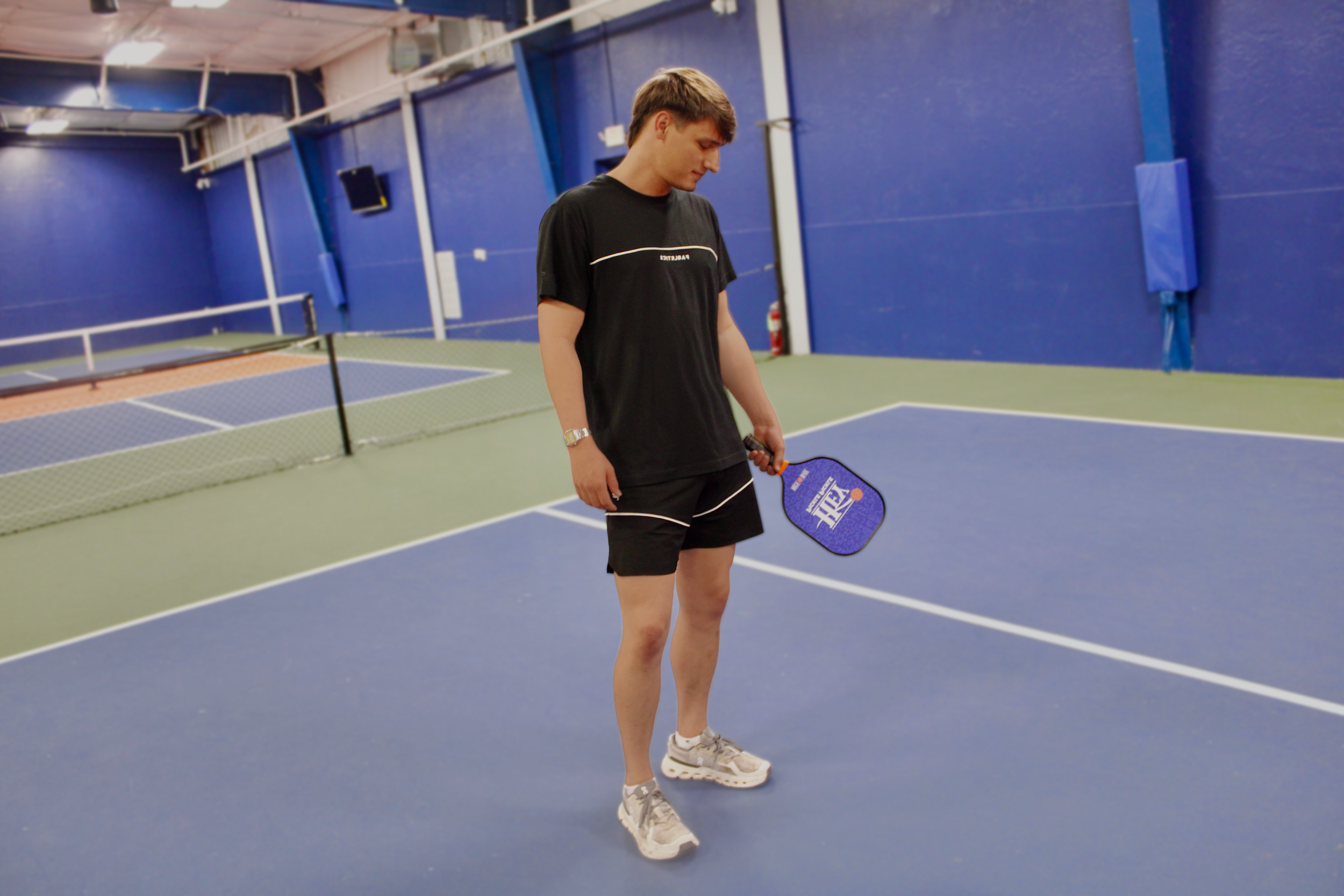 A young man standing on a blue indoor tennis court holding a tennis racket, wearing a black t-shirt, black shorts, and white sneakers, with a blue wall and tennis net in the background.