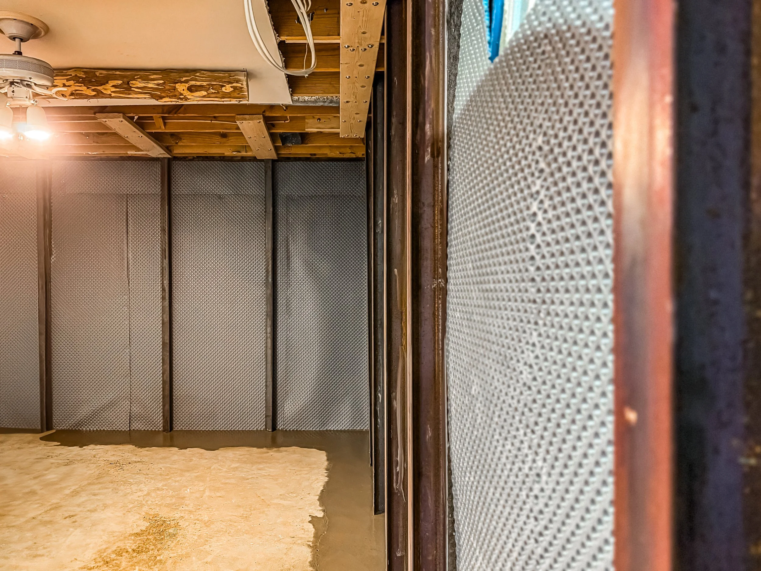 A construction site shows a partially finished wall with exposed wooden framing and insulation. The floor is unfinished and a work surface is on the right side of the image.
