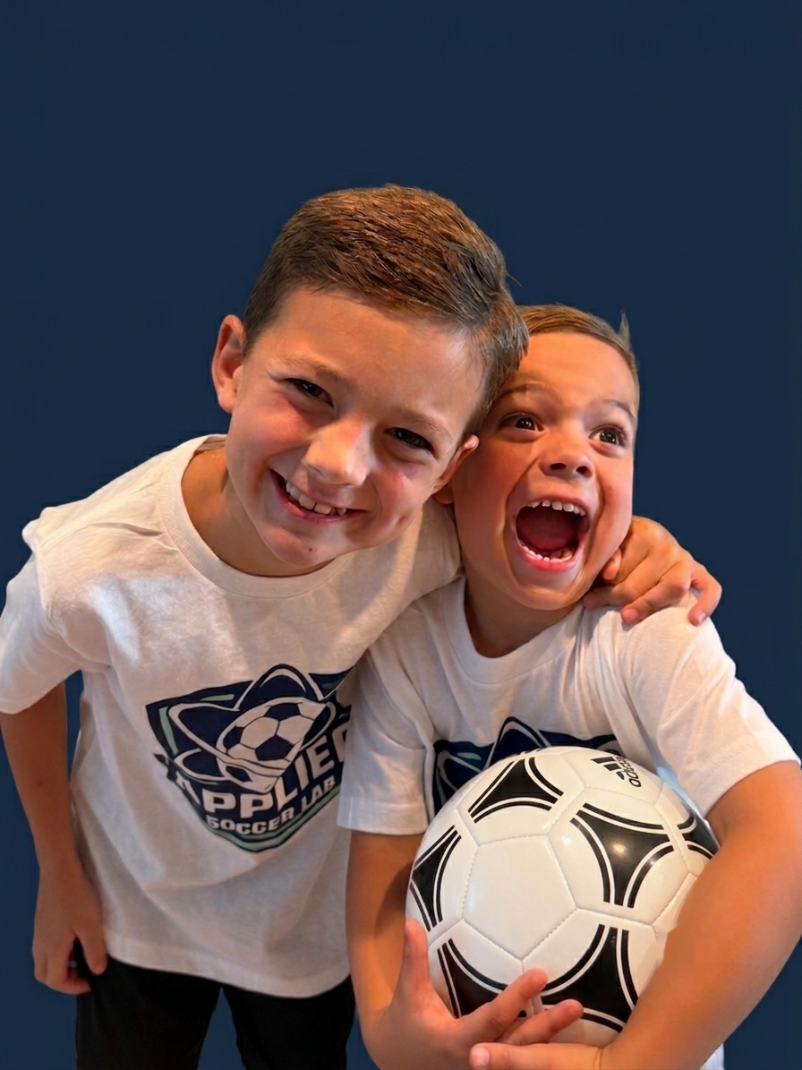 Two young boys in soccer shirts smiling, one holding a soccer ball, against a dark blue background.