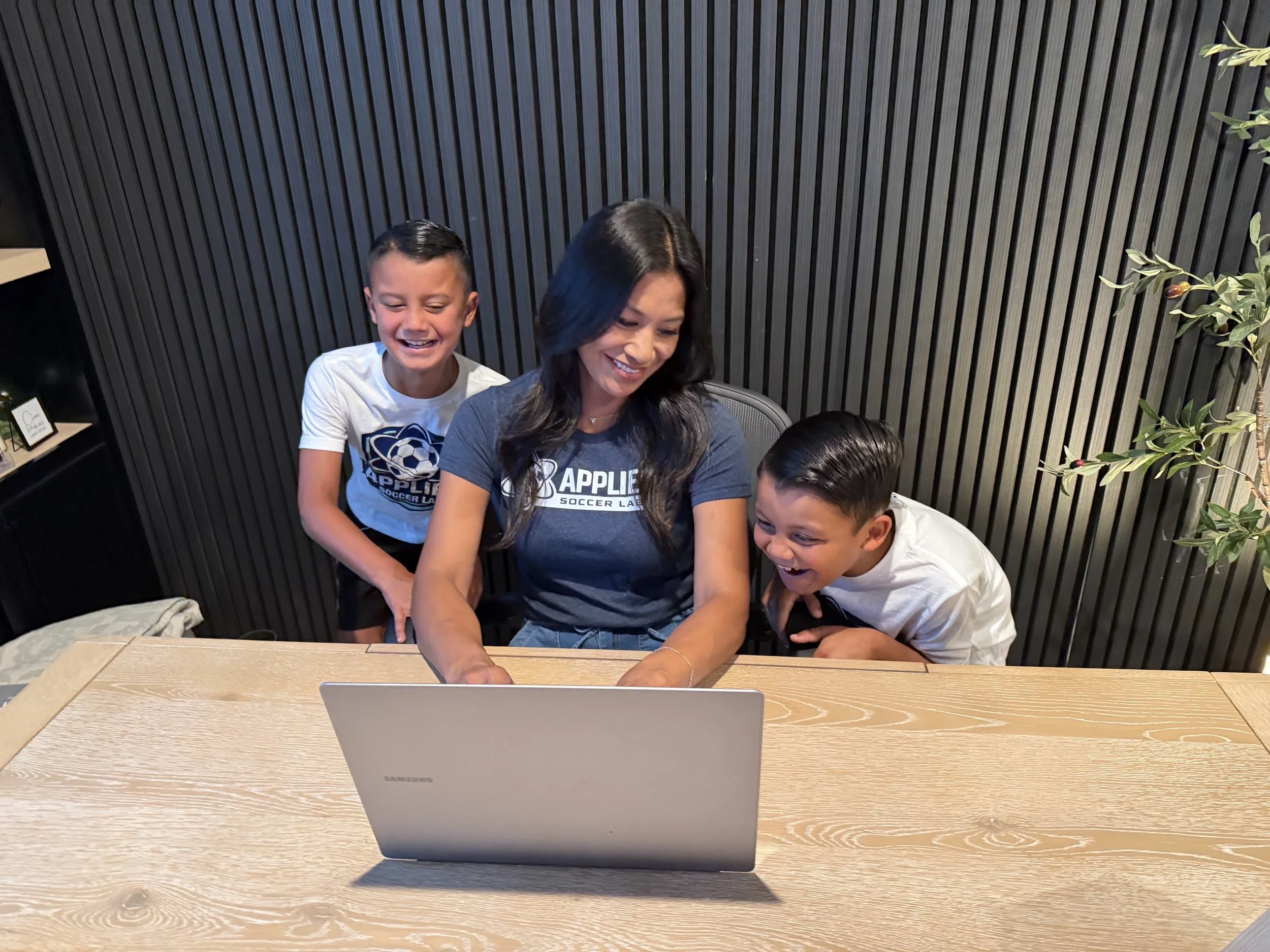 A woman and two young boys sitting at a wooden table, looking at a laptop screen and smiling. The woman is wearing a blue shirt with the logo 'APPLIE' and the boys are wearing white shirts. The background features a black slatted wall and a green plant.