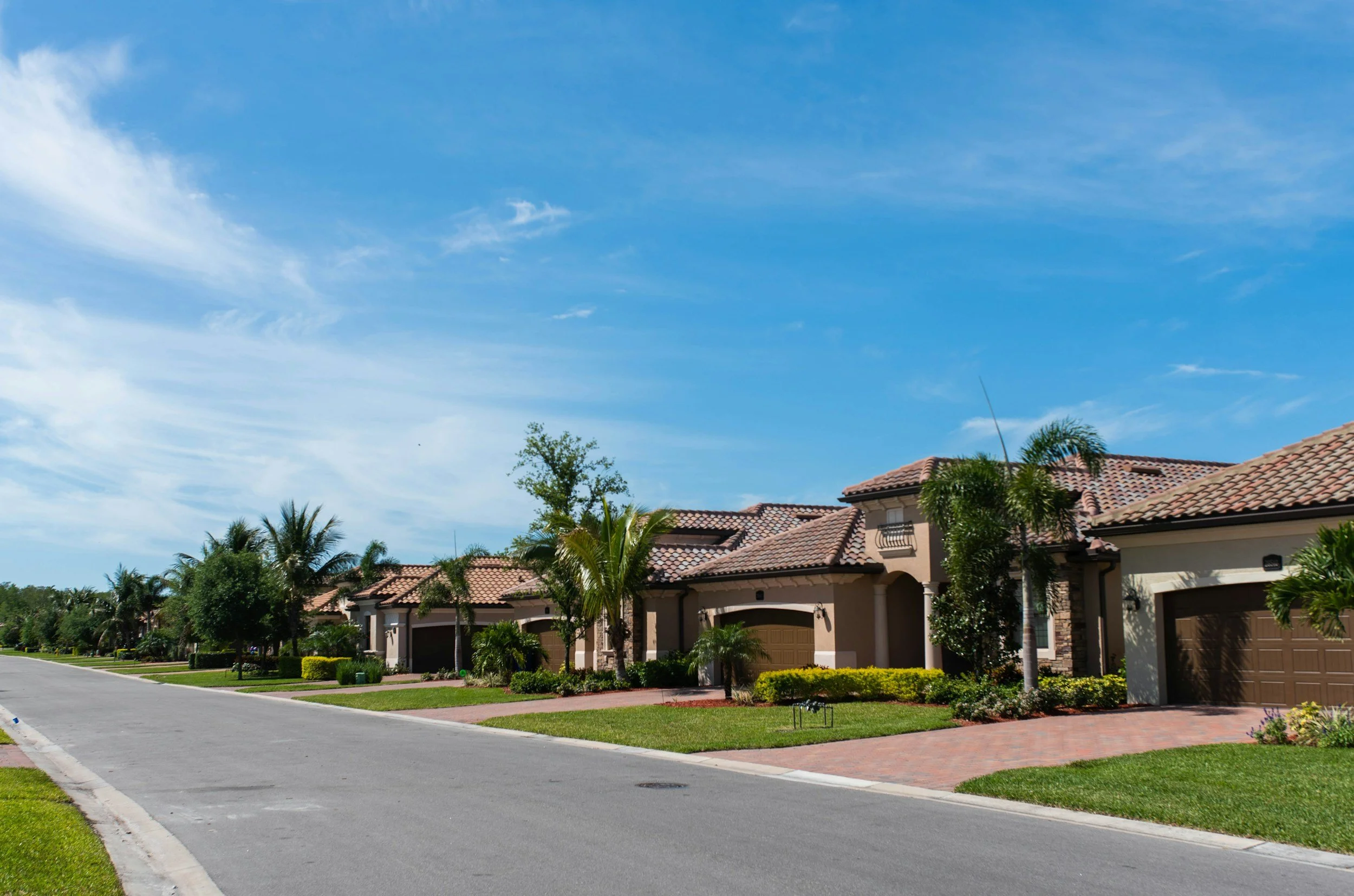 A suburban neighborhood street with row of houses featuring tiled roofs, palm trees, manicured lawns, and a clear blue sky with some clouds.