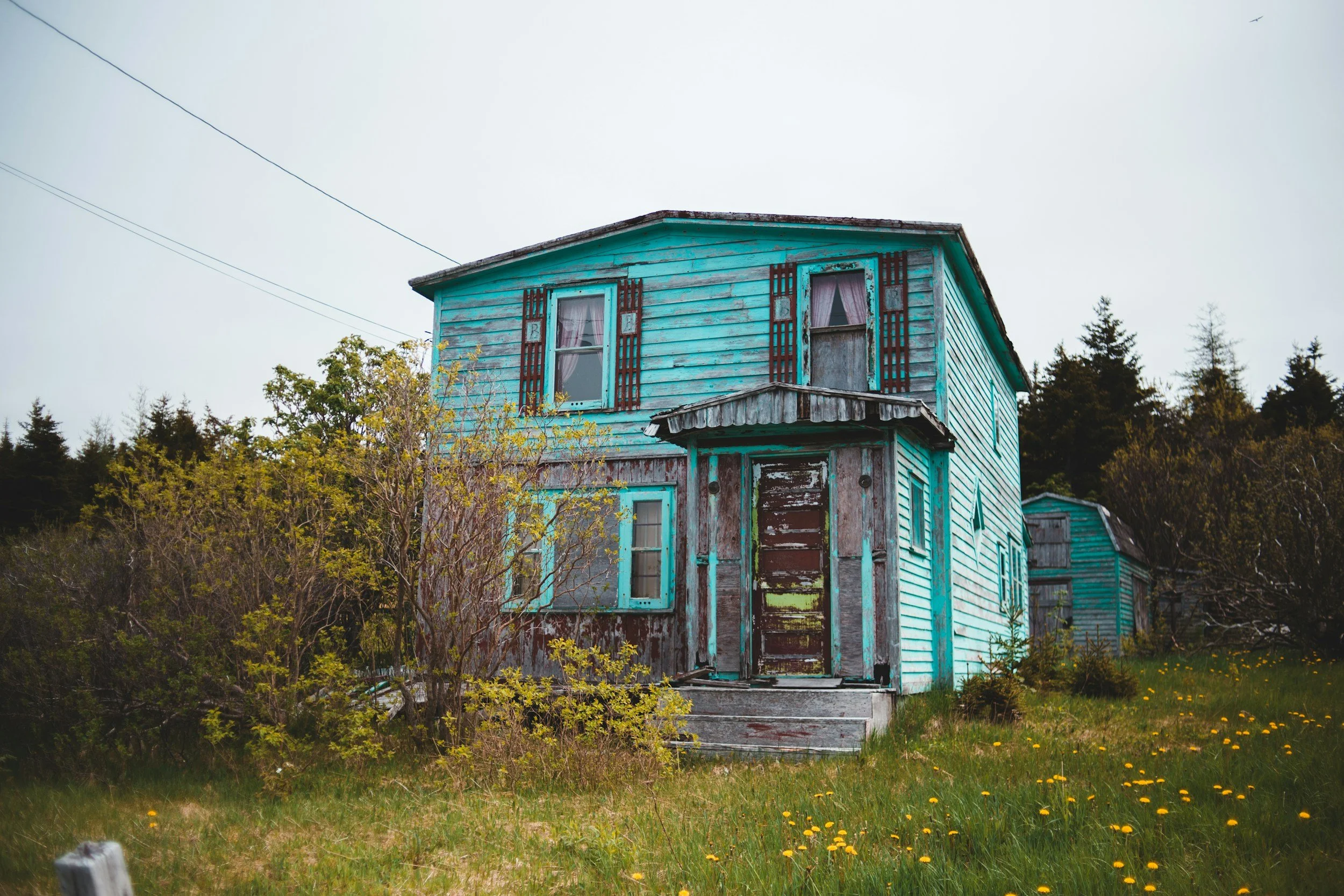 An old, weathered blue house with peeling paint and boarded-up windows, situated on a grassy lot with trees and a fence in the foreground.