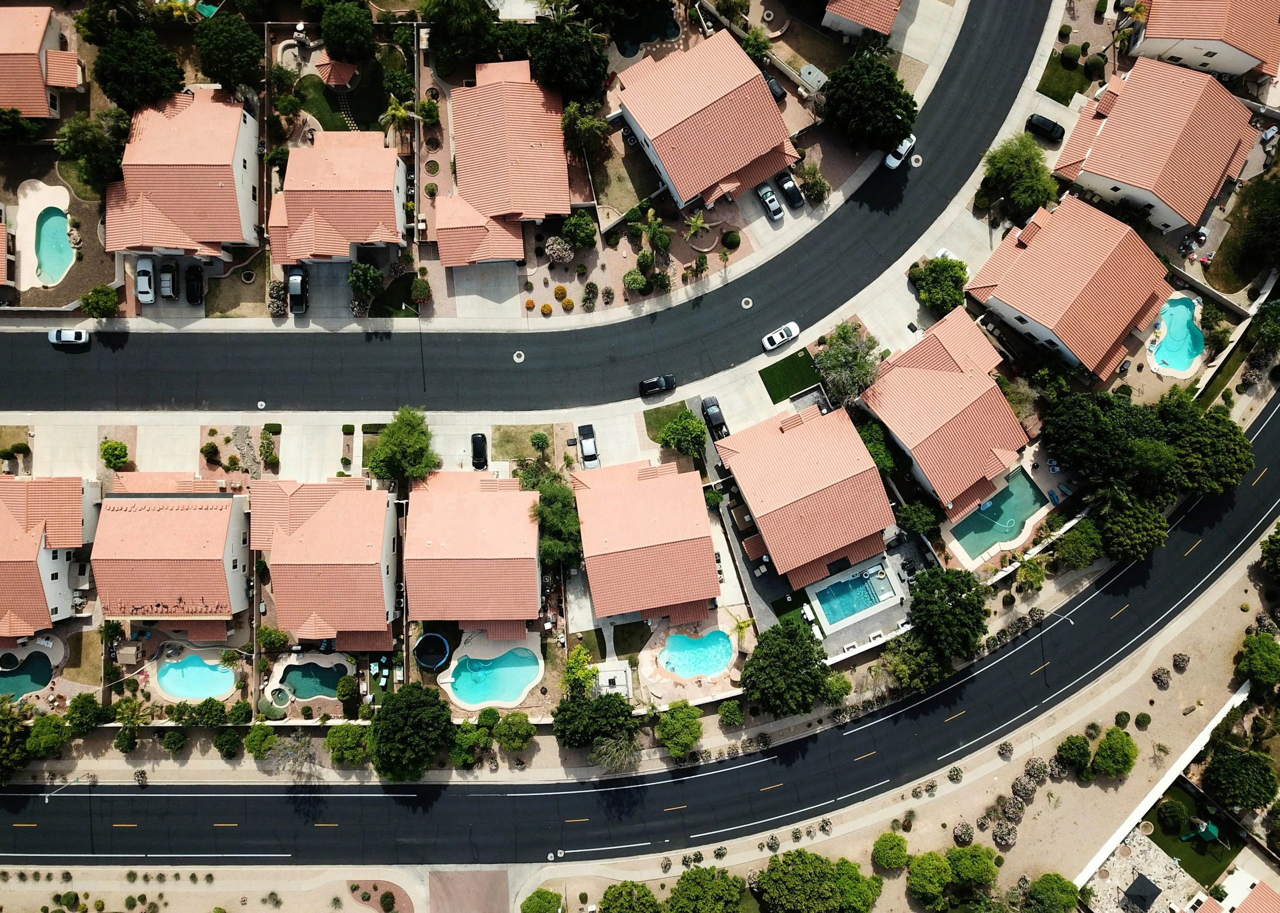 Aerial view of a suburban neighborhood with houses featuring red tile roofs, green trees, backyard swimming pools, and a curving main street.