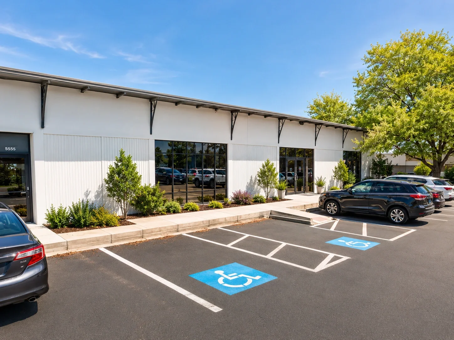 Parking lot with handicapped designated spaces, a modern building with large glass windows, some cars parked, green trees and shrubs, blue sky.