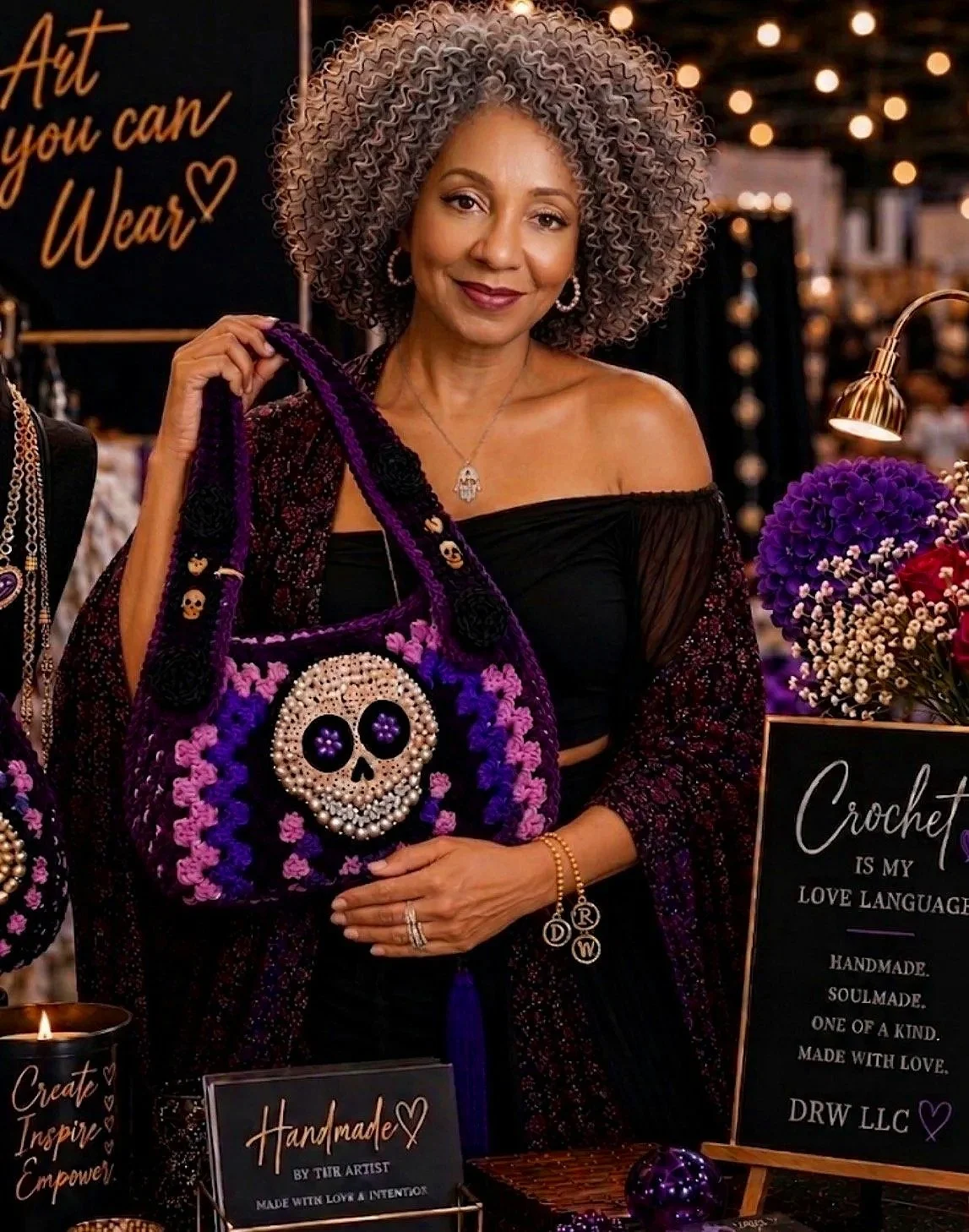 A woman with curly gray hair smiling at a craft booth, holding a purple and black crocheted bag with a skull design, surrounded by flowers and craft signs.