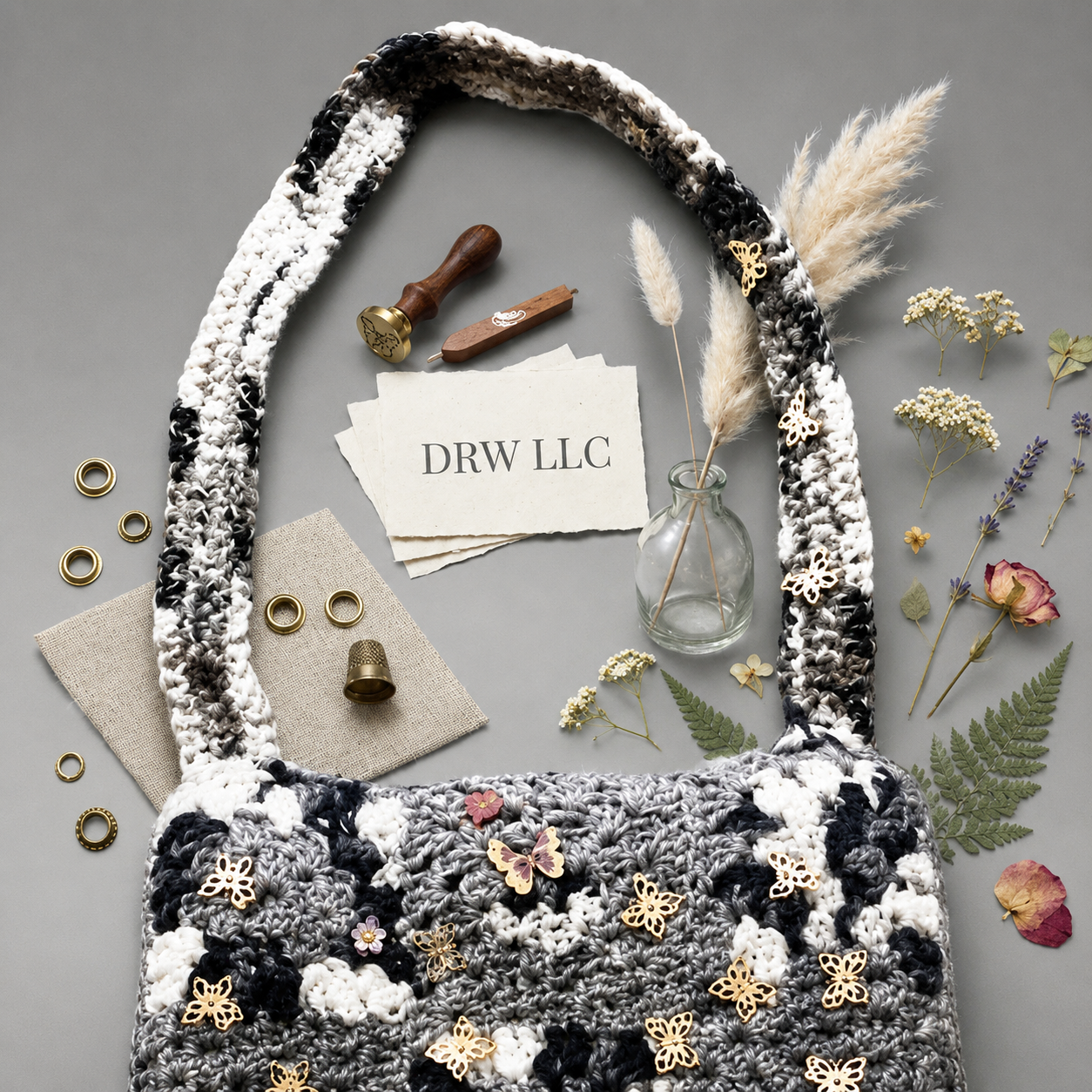A crochet bag decorated with butterfly pins, surrounded by dried flowers, gold rings, a small glass jar with pampas grass, and handwritten notes from DRW LLC, on a gray surface.