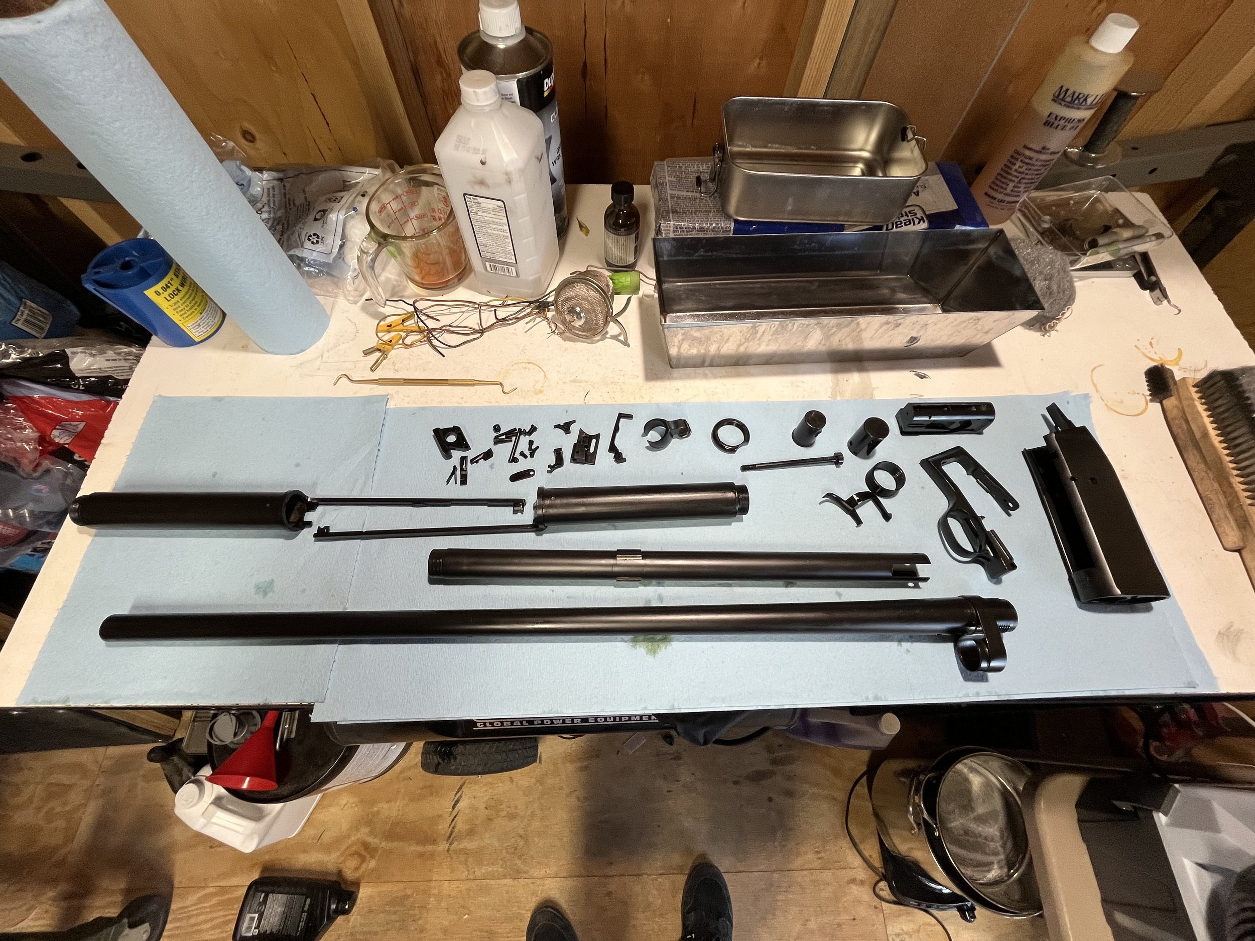 Disassembled motorcycle or bike parts on a workbench, including fork tubes, various small hardware, a motor, and tools, with a blue paper towel roll and lubricant bottles in the background.