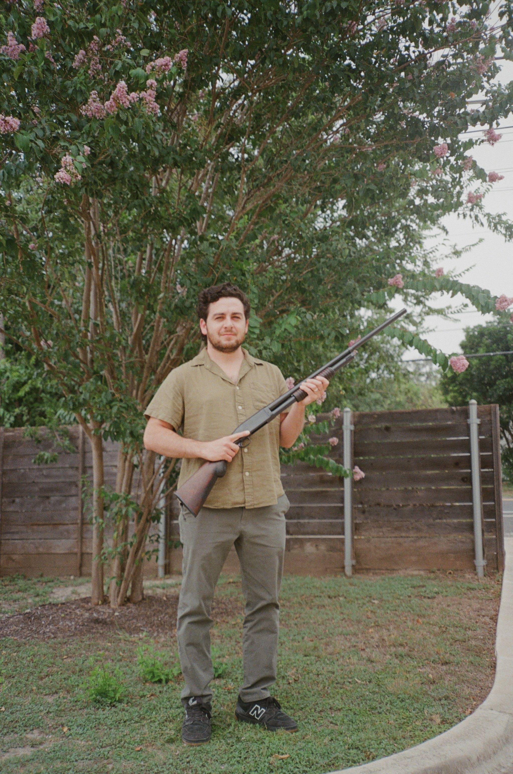 A young man with a beard and curly hair standing outdoors in front of a large tree with pink flowers, holding a rifle, wearing a beige button-up shirt, gray pants, and black sneakers.