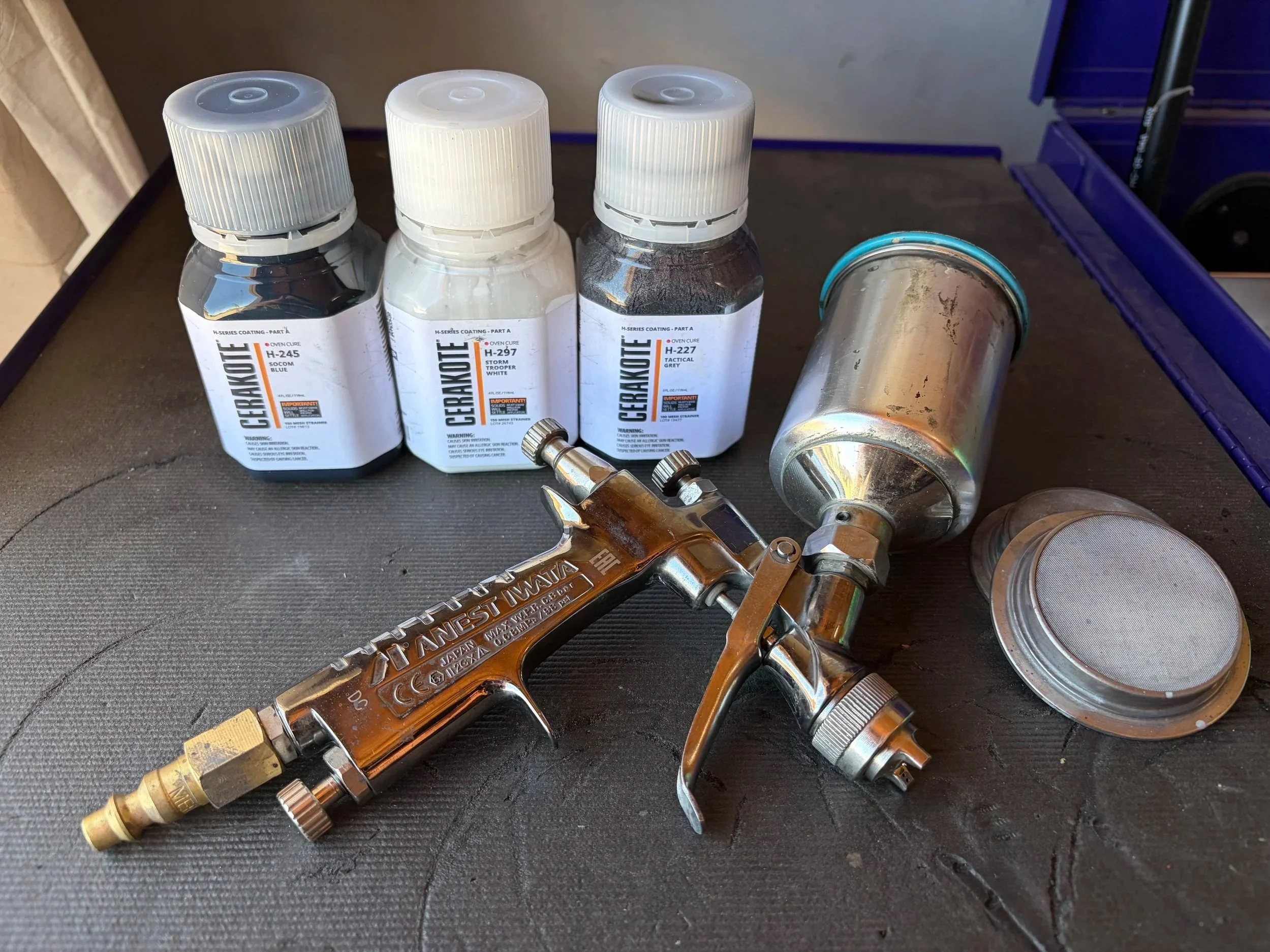 Three small bottles of ceramic coating with white caps, a spray gun, and a metal can, on a black textured surface.