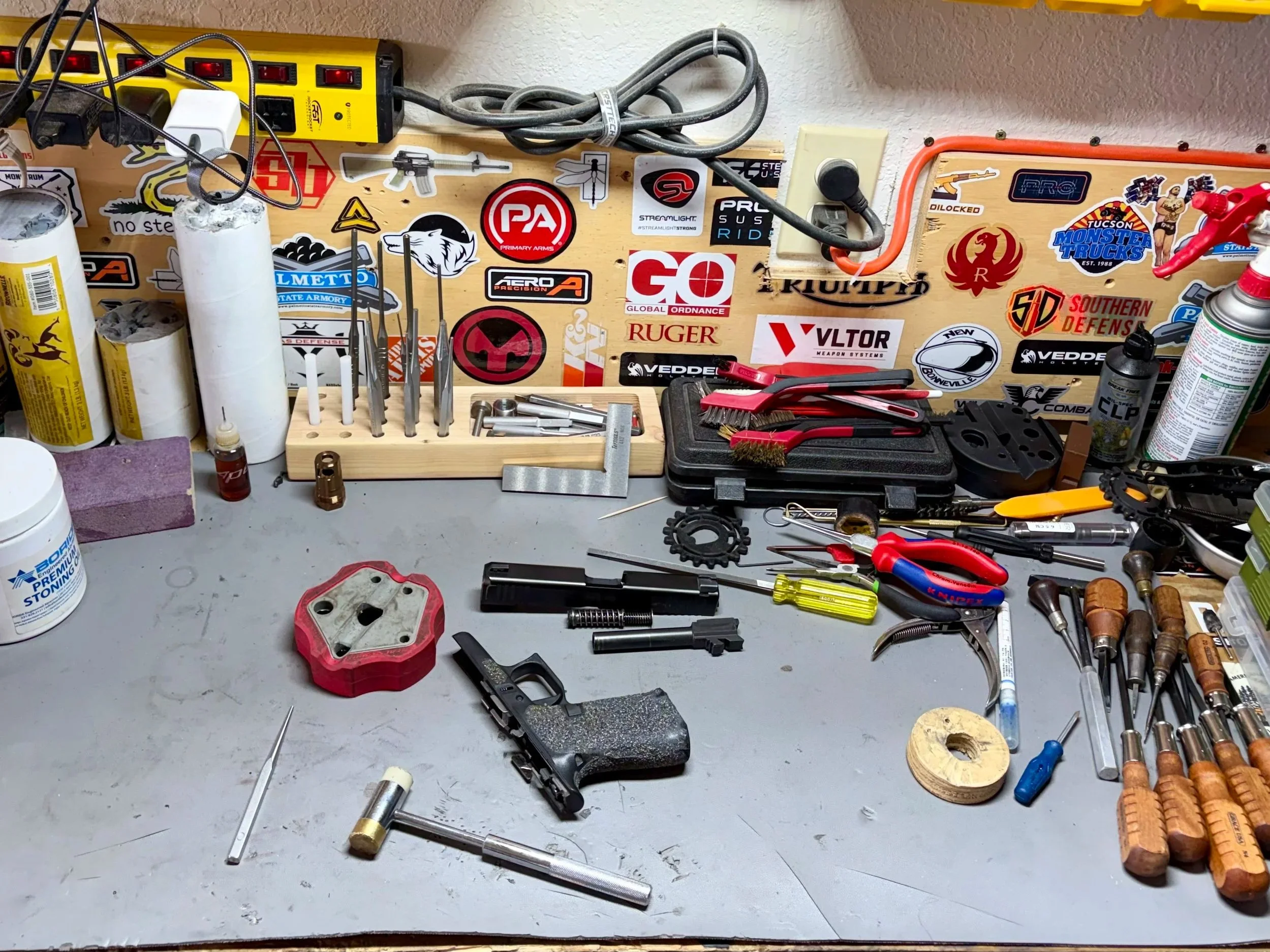 Workstation table with various tools, screws, and stickers on a wooden board mounted on the wall, including a power strip, wires, screwdrivers, and miscellaneous hardware.