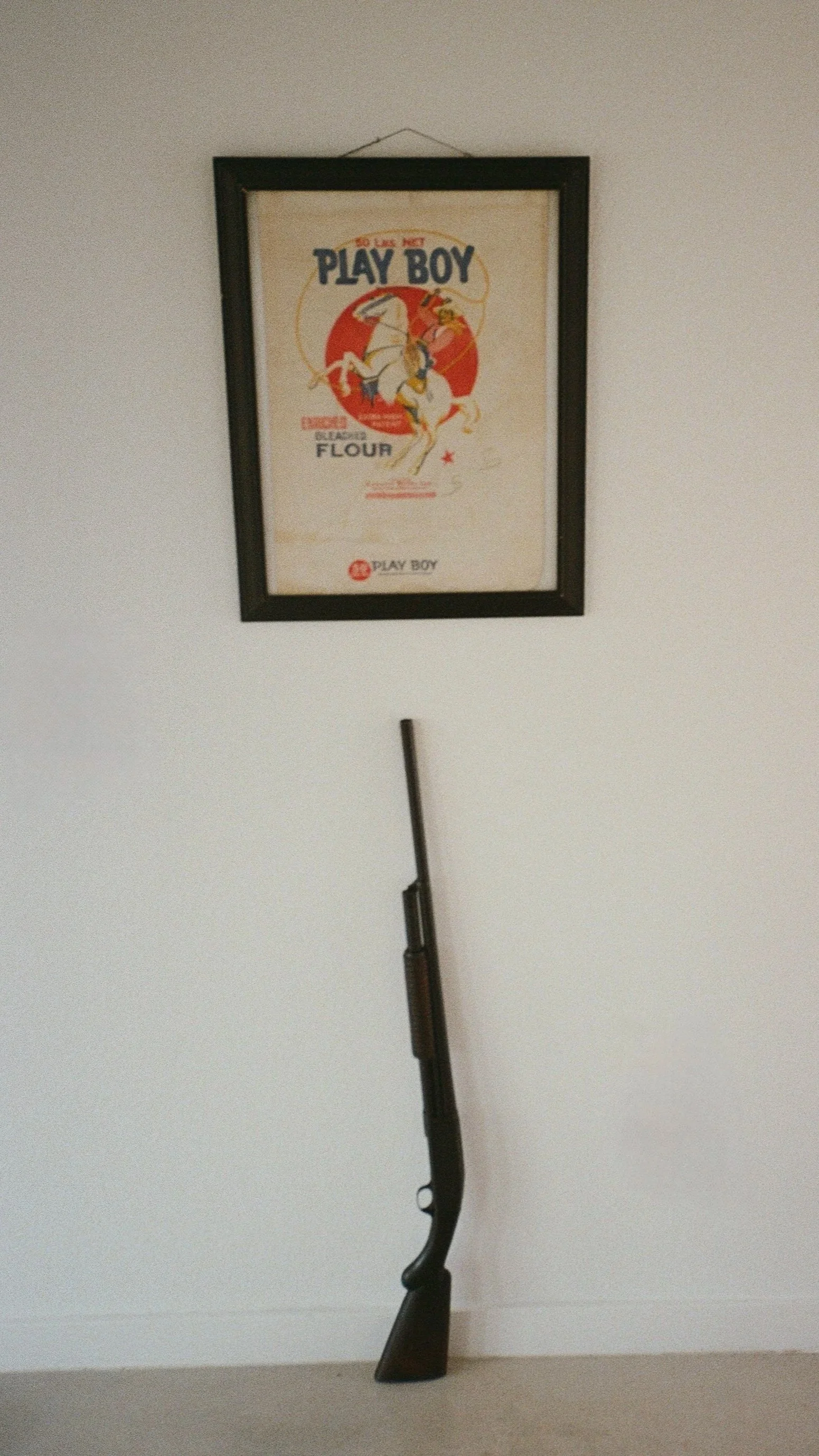 A framed vintage poster on the wall depicting a white horse and a person, with a gun leaning against the wall below.