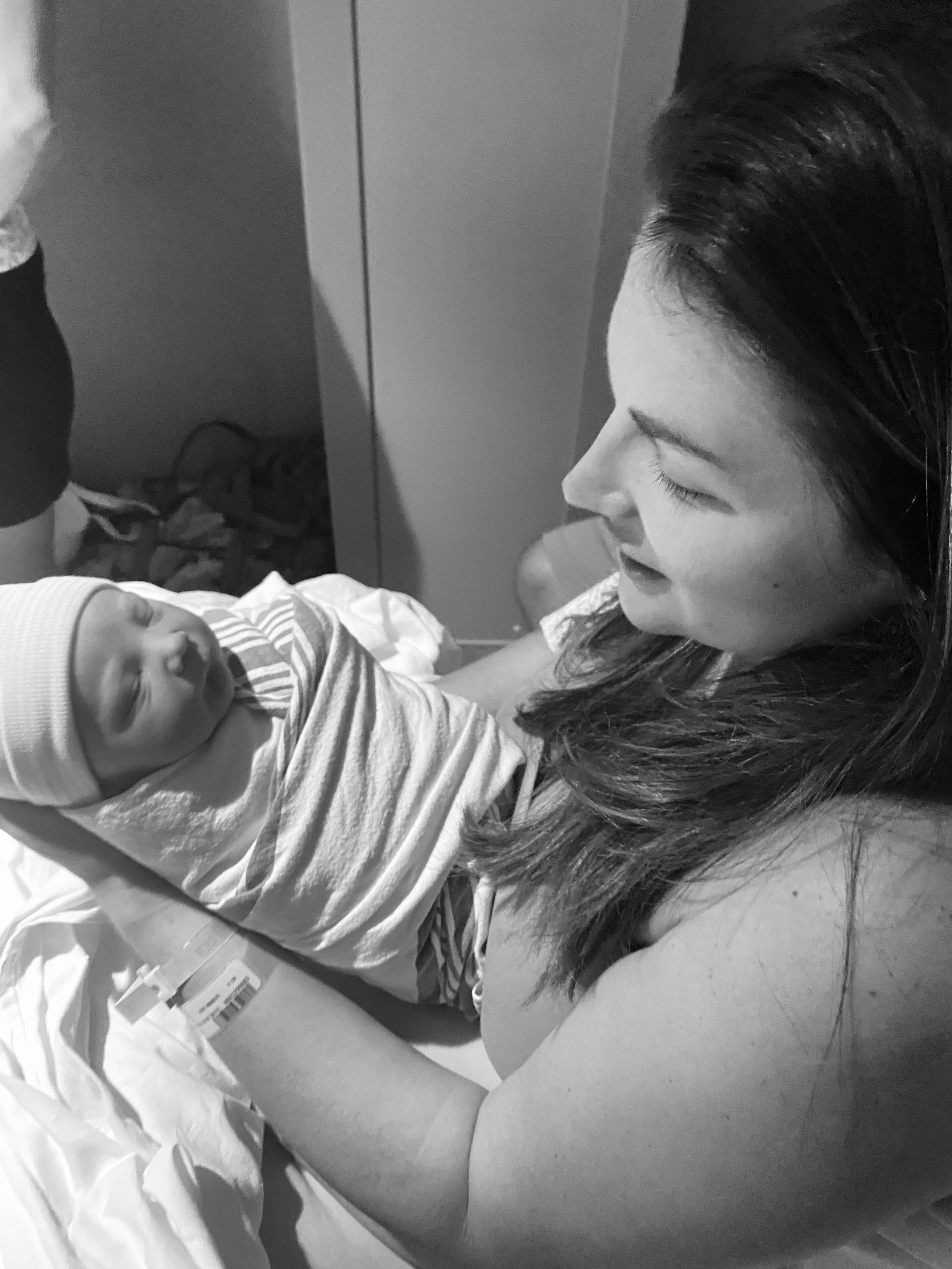 A woman with long dark hair tenderly looks at a newborn baby wrapped in a striped blanket, lying on a hospital bed with medical supplies nearby.
