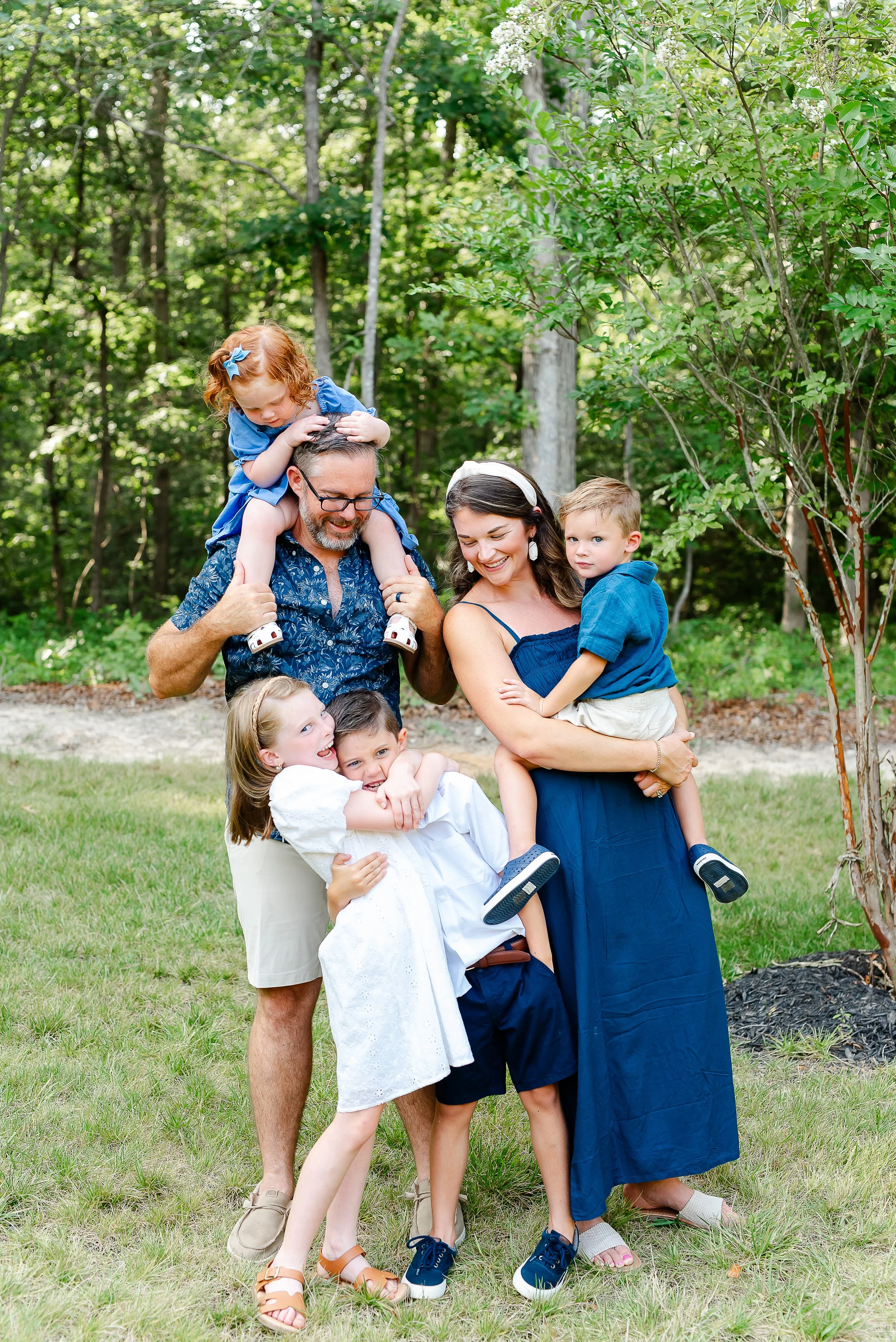 A family of five children and two adults enjoying a moment outdoors in a lush green park with trees and grass. The children are playing, and one child is being held by the mother. The father has a child on his shoulders, and all are smiling or looking happy.
