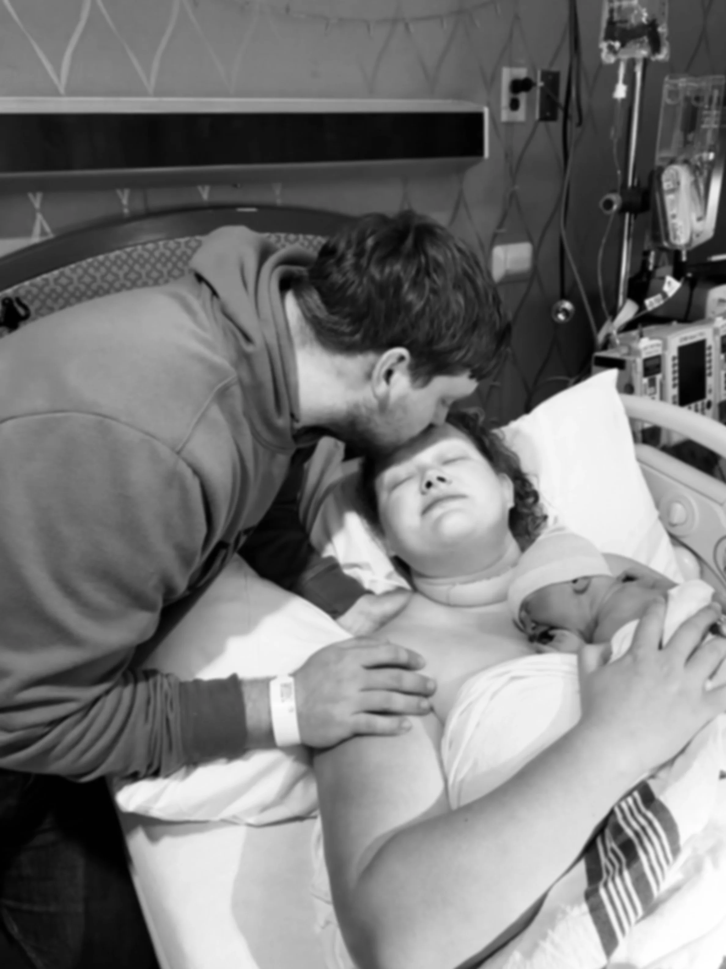 A man leaning over a woman lying in a hospital bed, holding her hand and kissing her forehead, with medical equipment and monitors nearby.